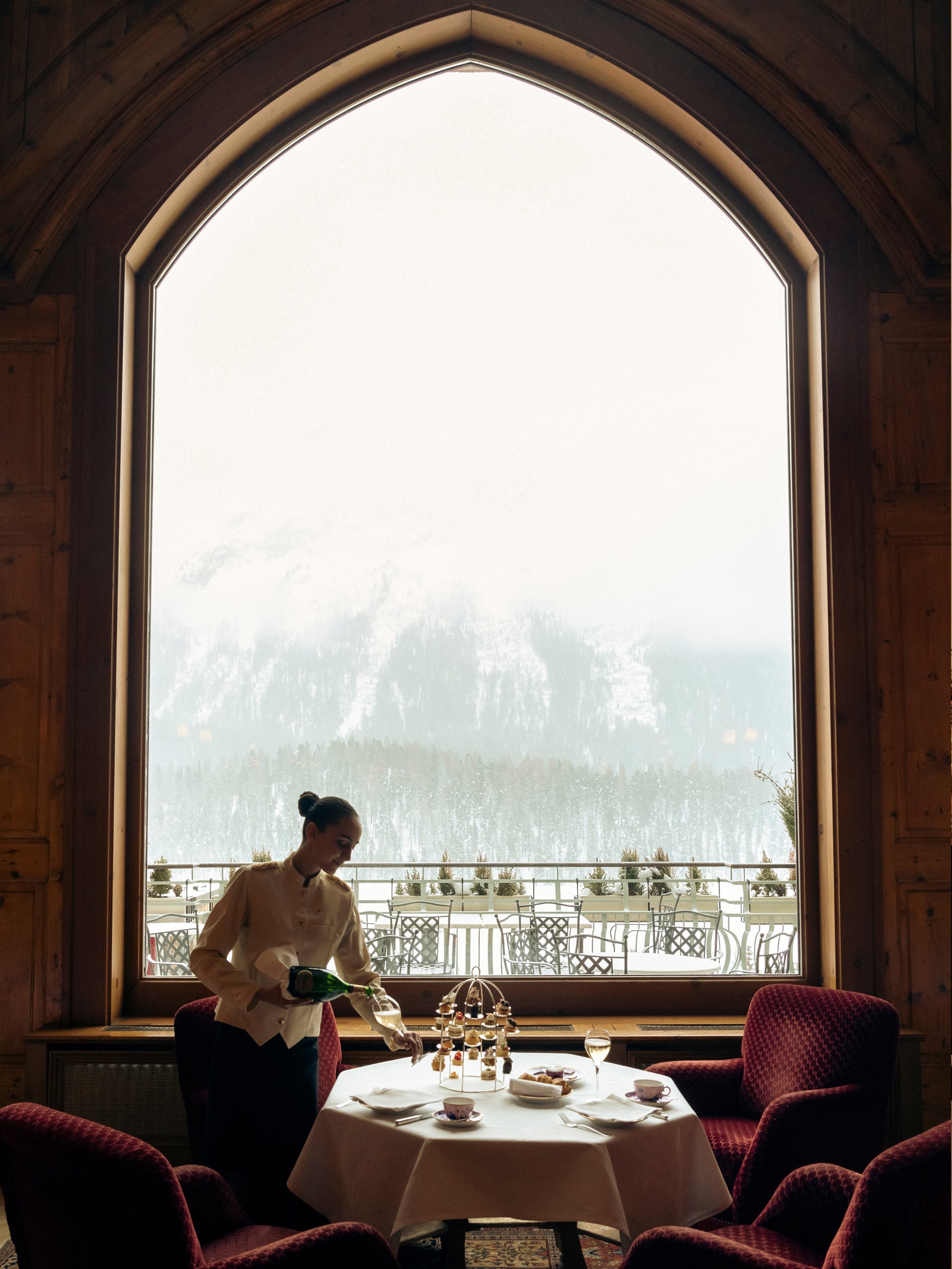 A server pours champagne beside an arched window framing snow-dusted Alpine peaks, where intimate dining unfolds against Switzerland's dramatic mountain theater.