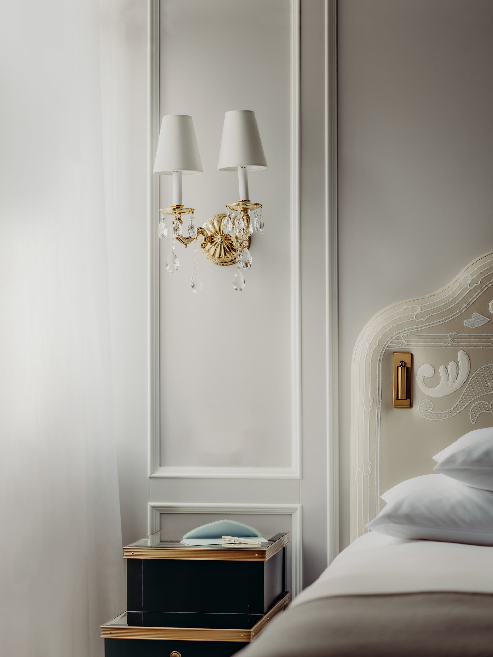 Crystal chandelier sconces cast warm light across ornate headboard details, where gilded fixtures and crisp white linens create an atmosphere of refined Alpine luxury.