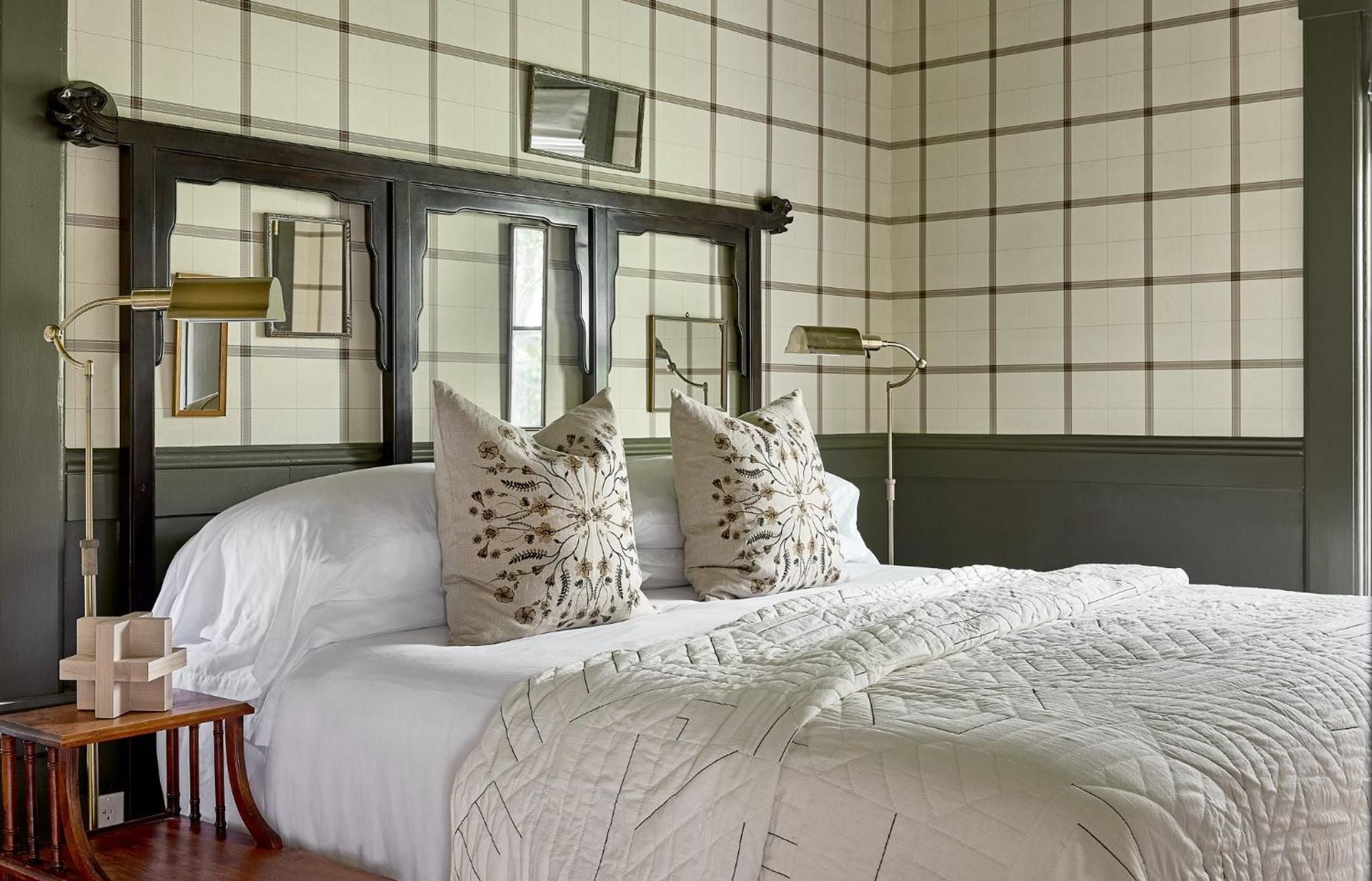 Plaid wallpaper and brass swing-arm sconces frame the mirrored headboard, while embroidered pillows add botanical charm to this thoughtfully appointed guest room.