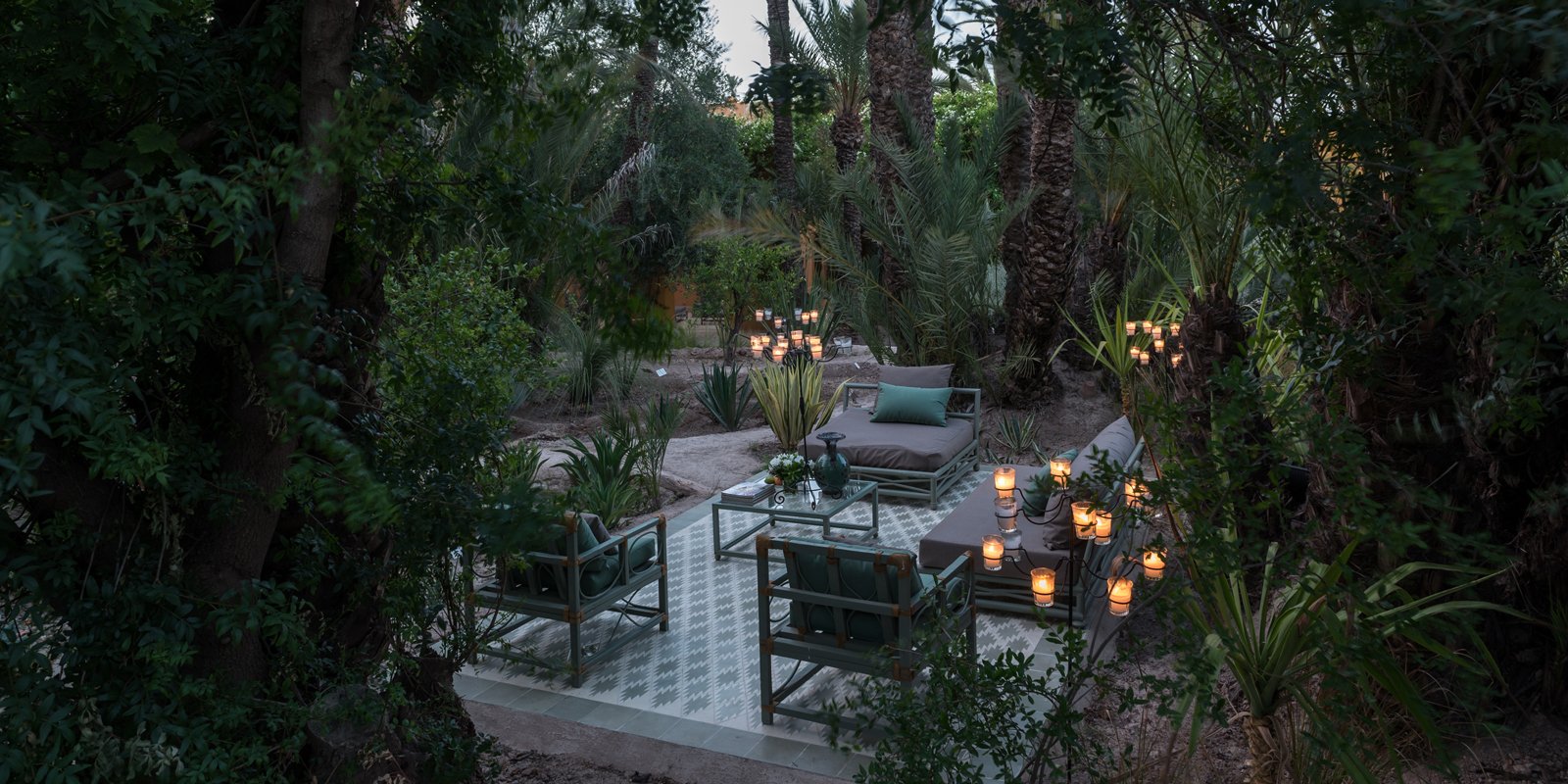 Candlelit terraces nestled among palms and desert flora create intimate outdoor sanctuaries where modern furniture meets Morocco's ancient oasis spirit at dusk.