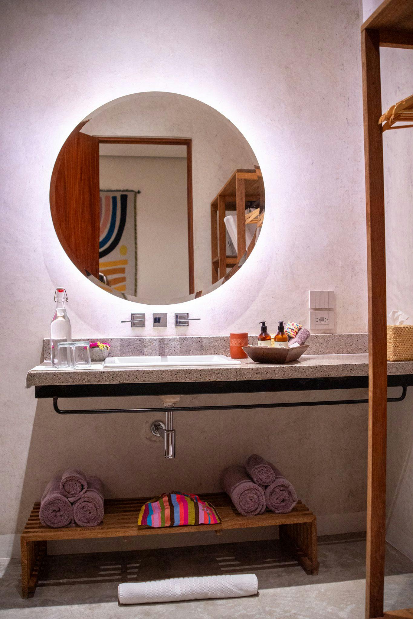 Warm terrazzo and rich wood create an inviting vanity where rolled towels await beneath a glowing circular mirror, blending modern luxury with thoughtful details.