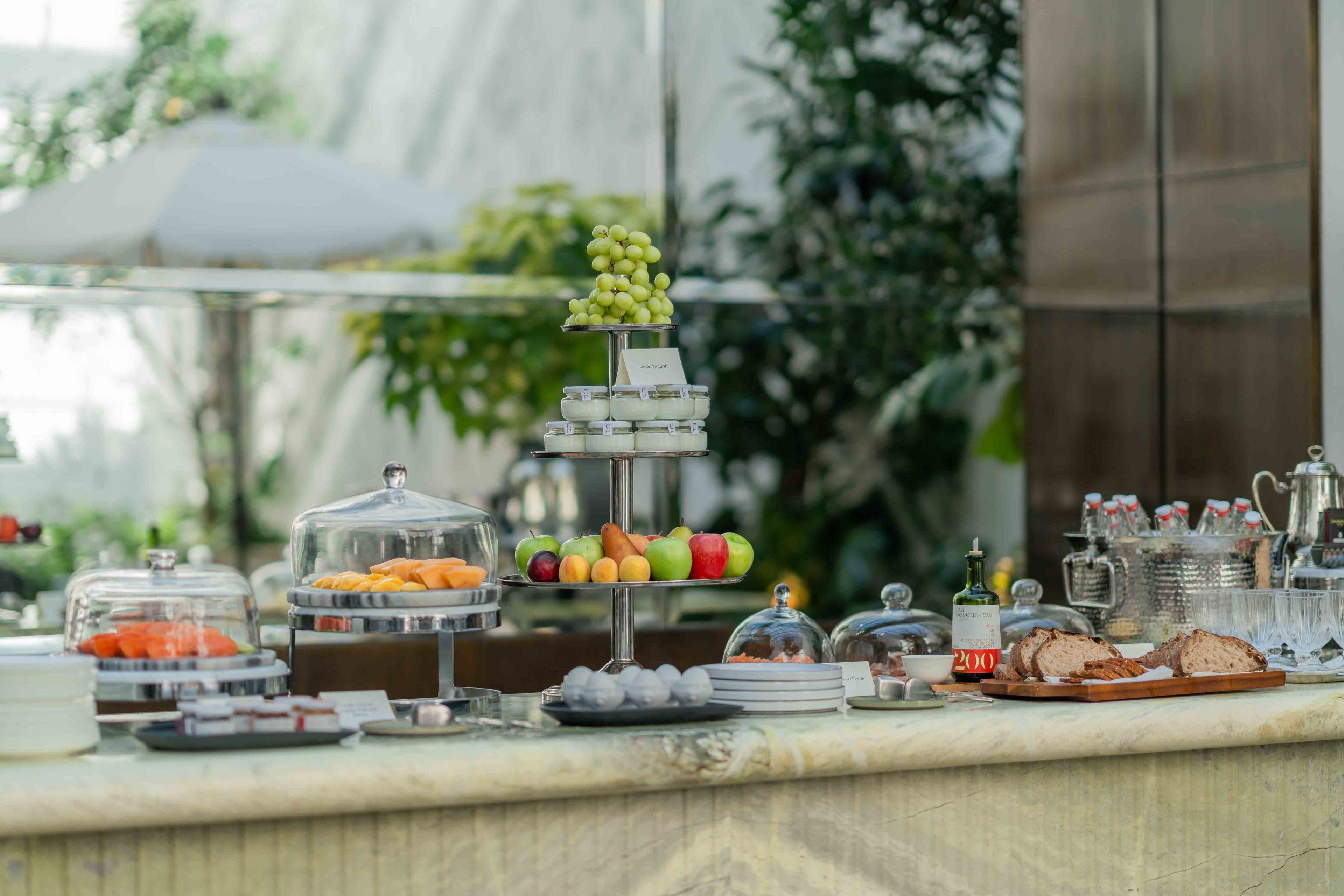 Marble countertops showcase an artful breakfast spread where fresh grapes crown tiered platters of seasonal fruit, beneath glass domes protecting delicate pastries in natural light.