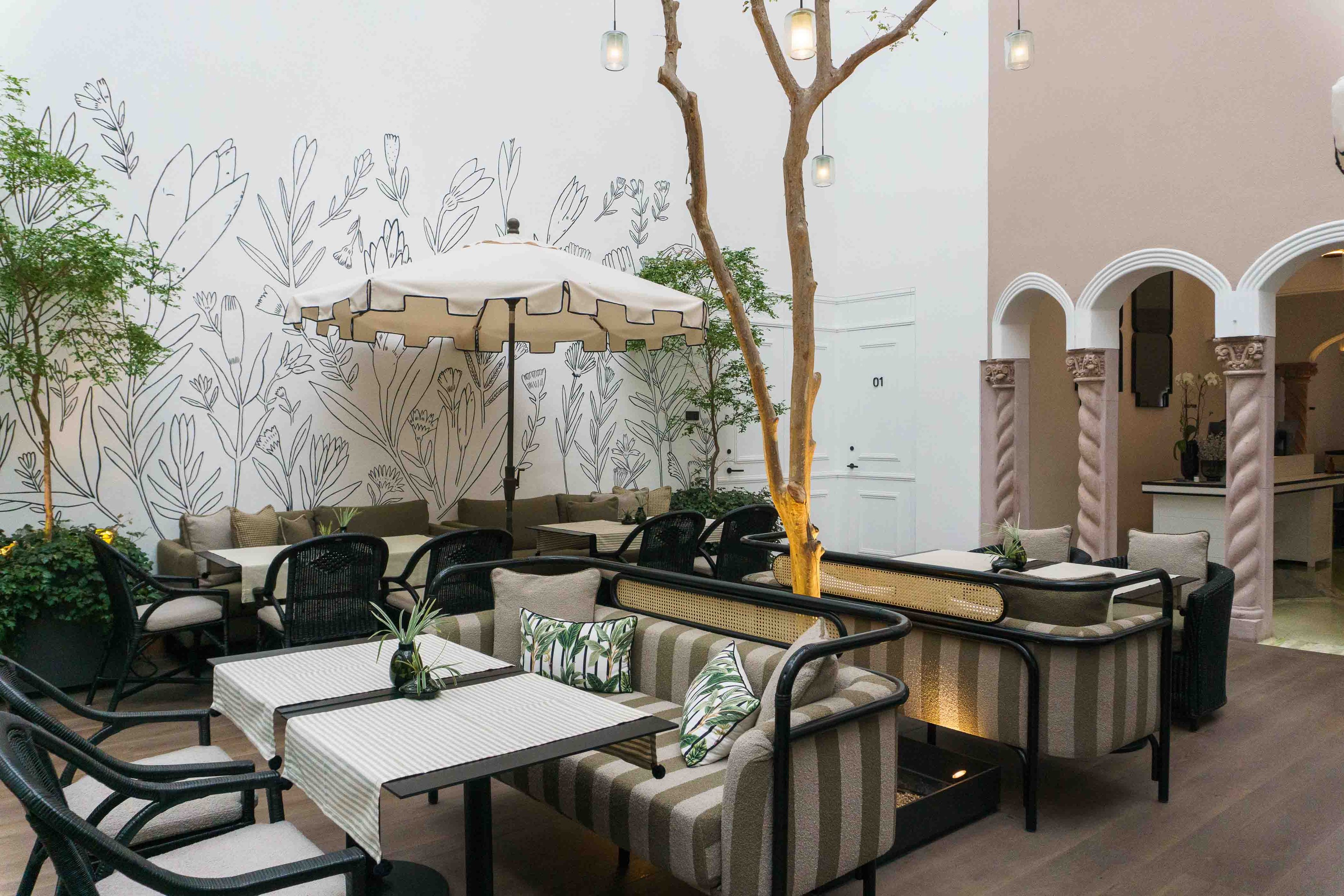 Hand-drawn botanical murals create an enchanting backdrop in the courtyard lounge, where rattan furniture and string lights foster intimate conversations beneath a living tree canopy.