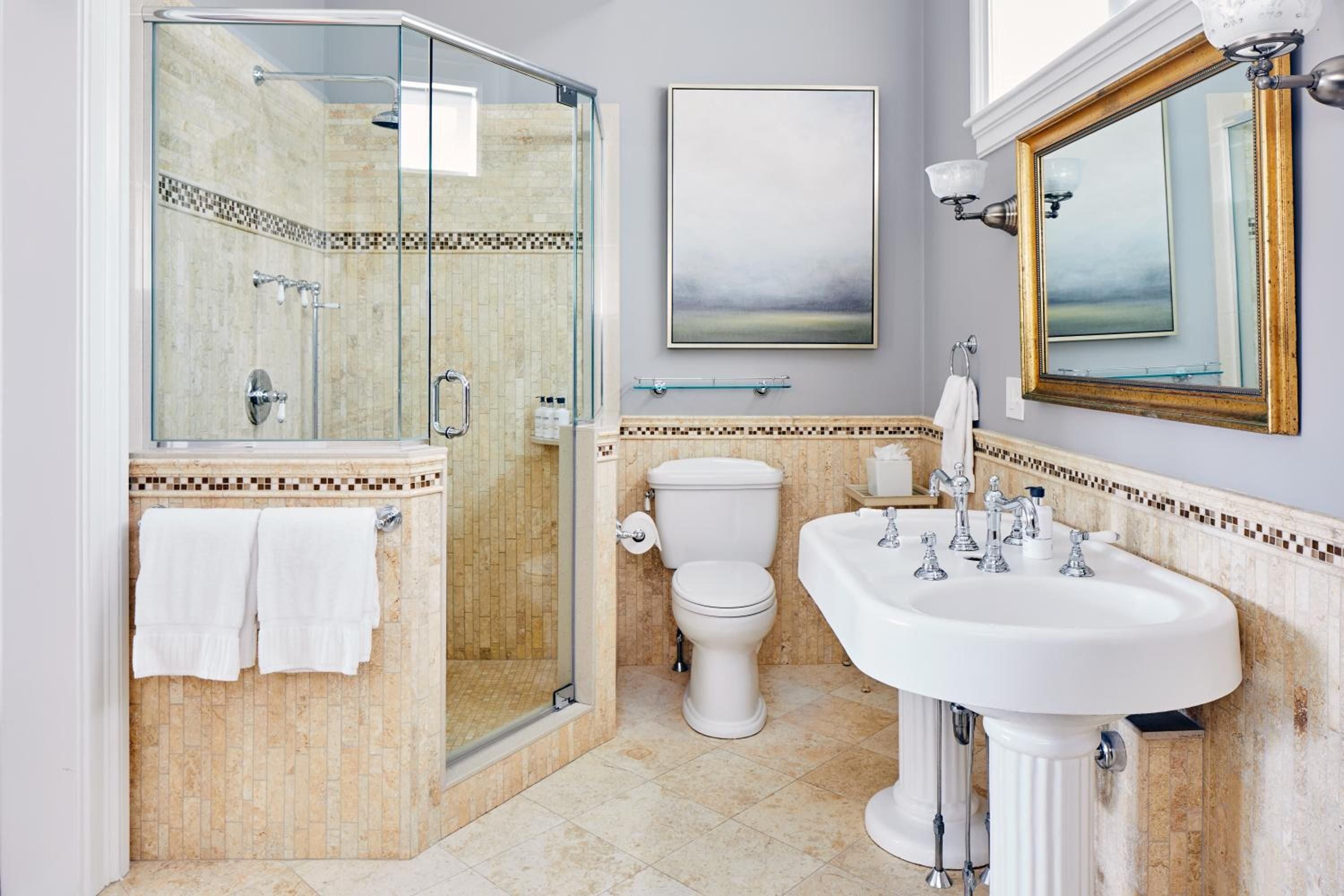 Mosaic tile borders and chrome fixtures create spa-like elegance in this thoughtfully appointed guest bathroom, where glass-enclosed showers meet classic pedestal sinks.