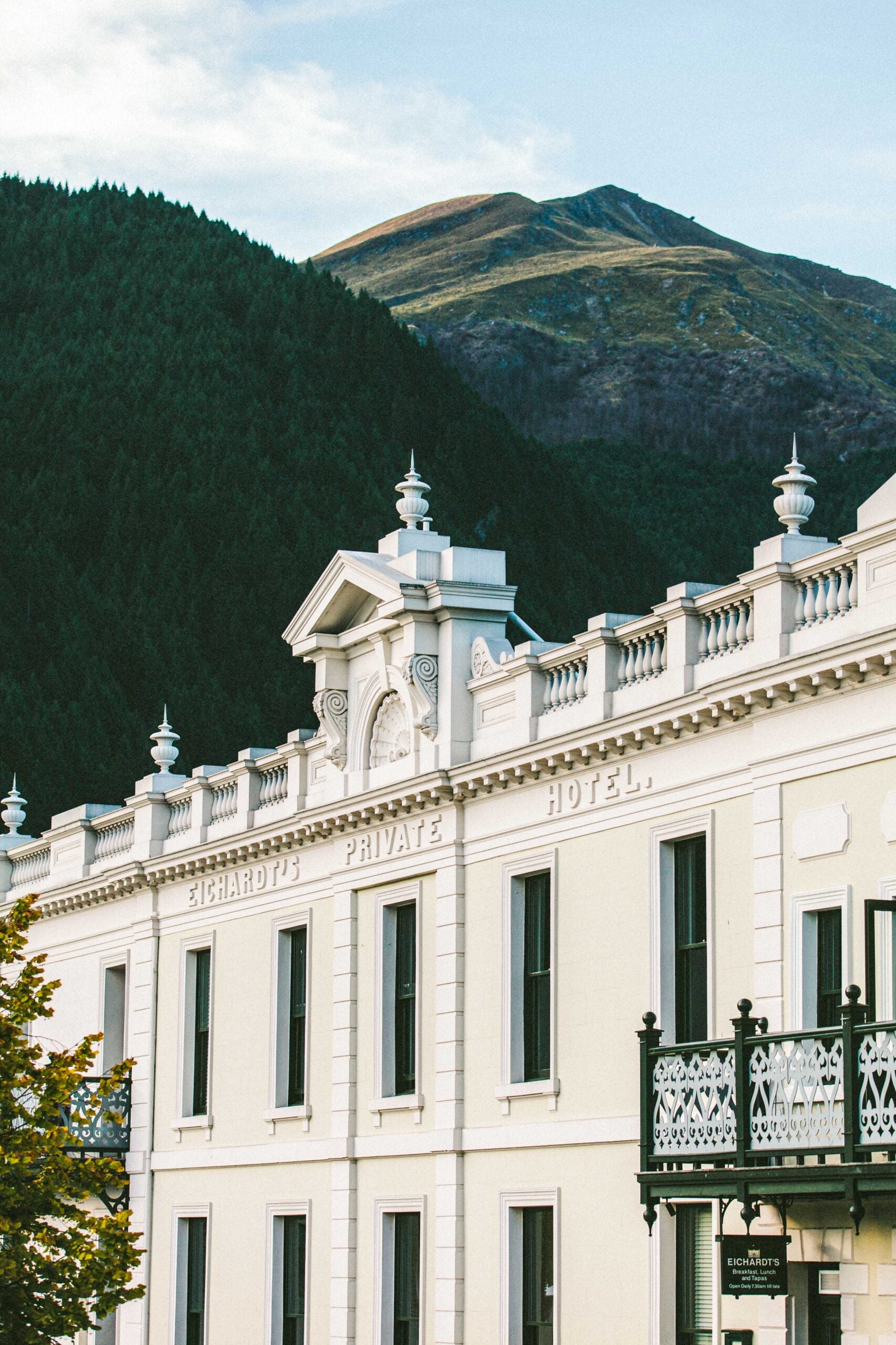 Victorian grandeur meets dramatic alpine backdrop as Eichardt's ornate white facade and decorative ironwork balcony frame the rugged mountains beyond Queenstown's lakeside setting.