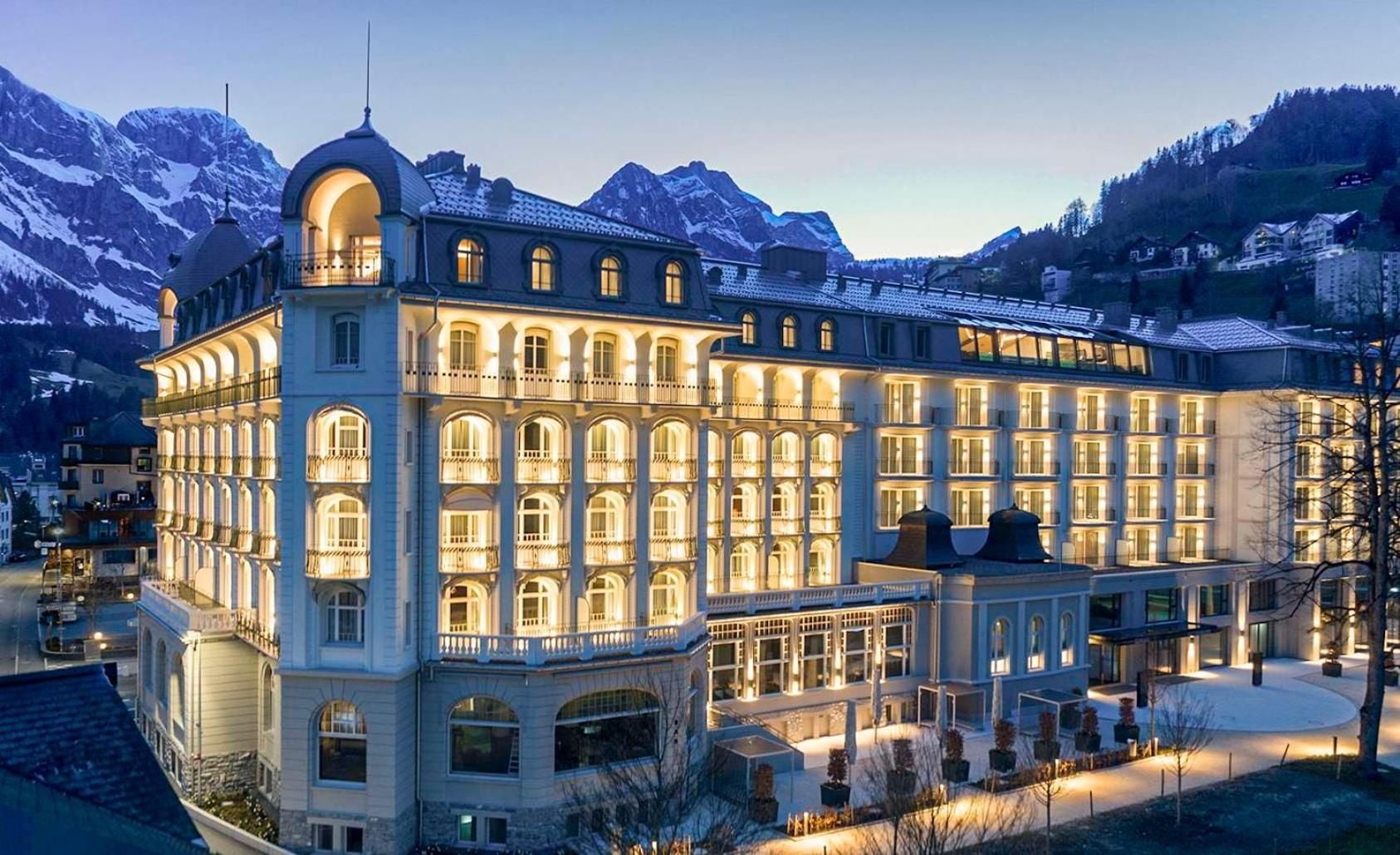 Illuminated against twilight skies, this grand Alpine palace glows warmly beneath snow-dusted peaks, its Belle Époque balconies and ornate facade promising old-world luxury in Engelberg's mountain embrace.