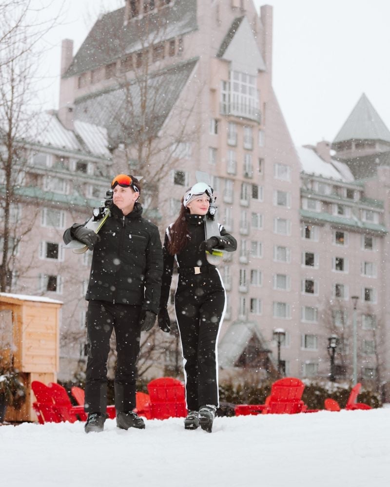 Fresh powder falls over the château's distinctive alpine architecture as guests embrace Whistler's winter magic, ski gear in hand beneath snow-laden eaves.