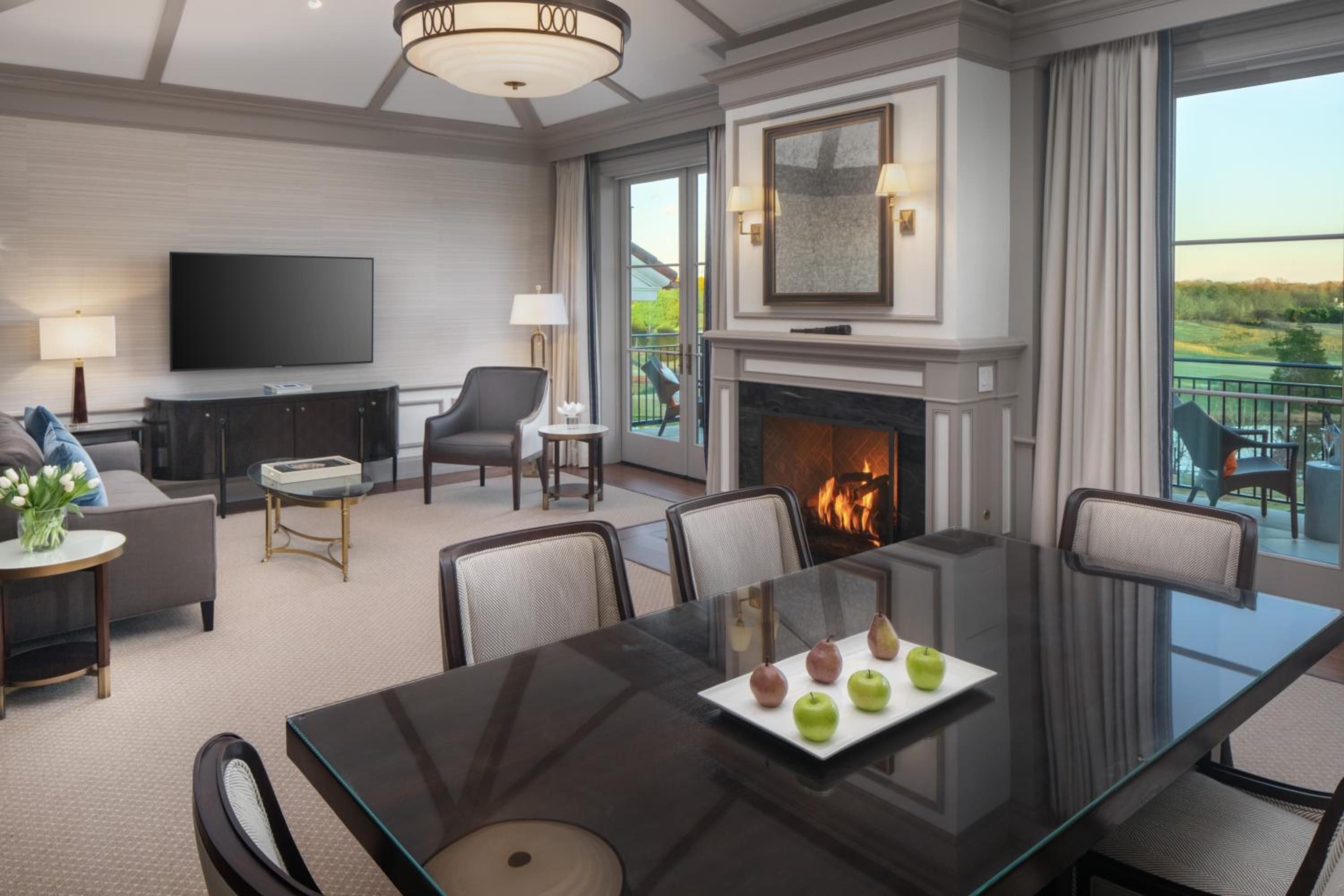Coffered ceilings and a crackling fireplace create intimate elegance in this spacious suite, where glass dining tables reflect golden flames and balcony doors frame rolling countryside views.