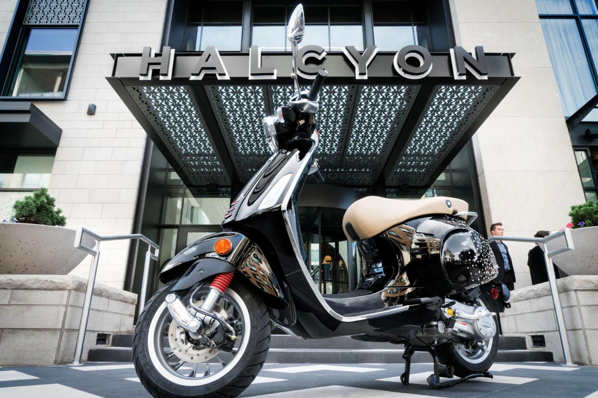 A gleaming vintage-style scooter awaits outside Halcyon's modern entrance, where polished chrome meets contemporary geometric canopies in perfect urban harmony.