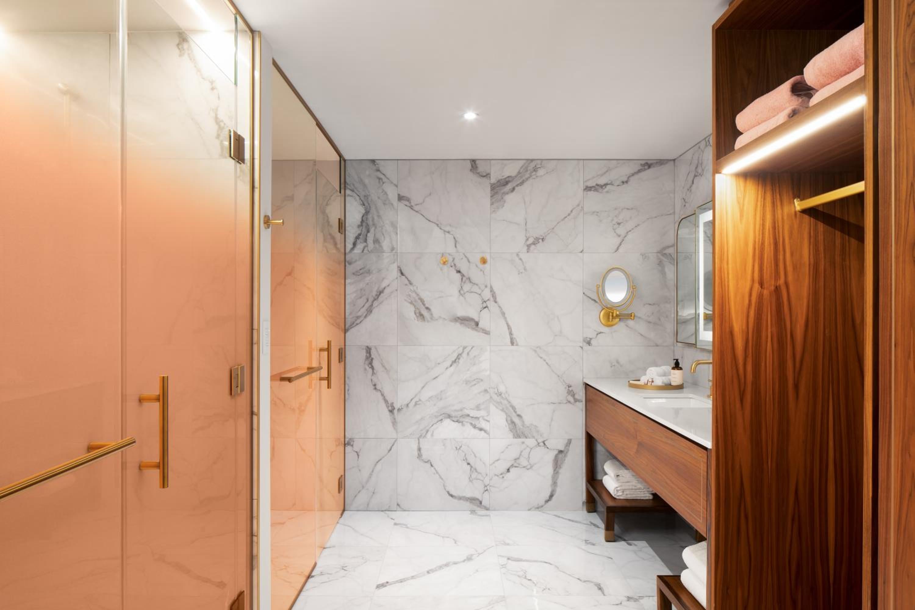 Calacatta marble walls meet warm walnut cabinetry in this striking bathroom, where brass fixtures and pink-toned glass create an unexpectedly glamorous sanctuary.