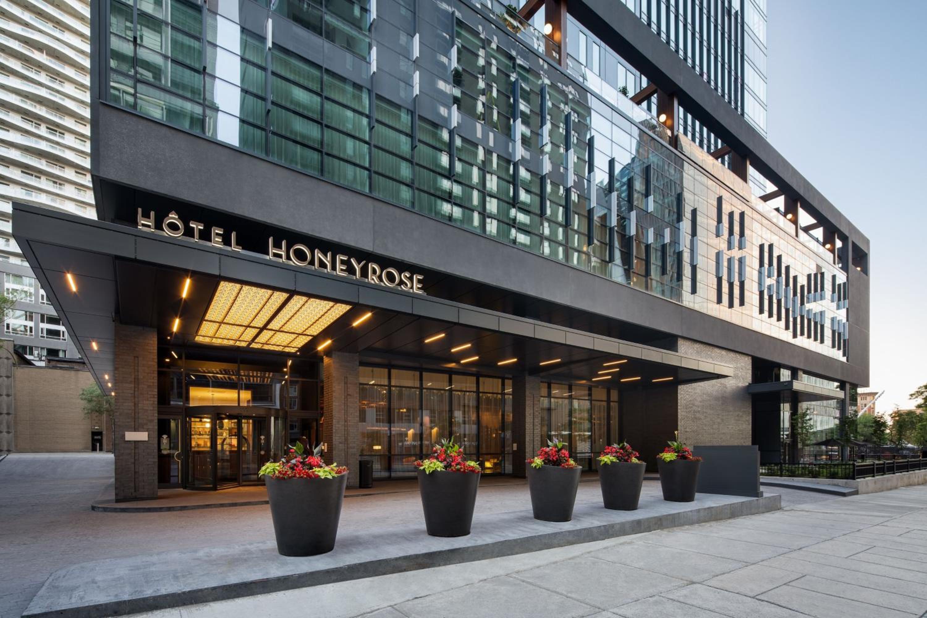 Sleek charcoal facades and warm amber lighting create an inviting urban sanctuary, where vibrant planters soften the contemporary glass-and-steel architecture against the evening sky.