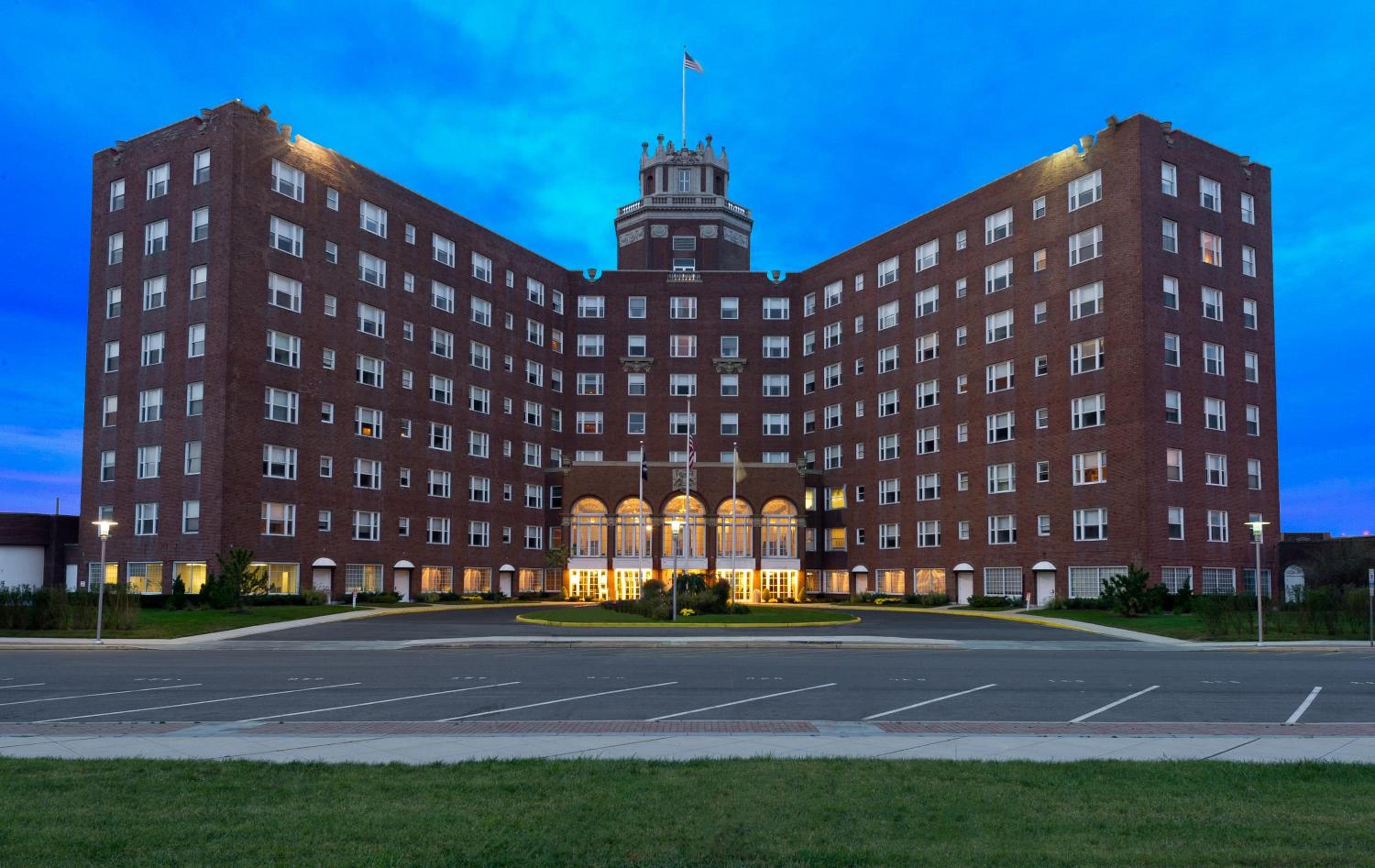 Stately brick wings embrace a grand illuminated entrance as twilight deepens, while the distinctive tower crowned with flags commands the evening skyline with old-world elegance.