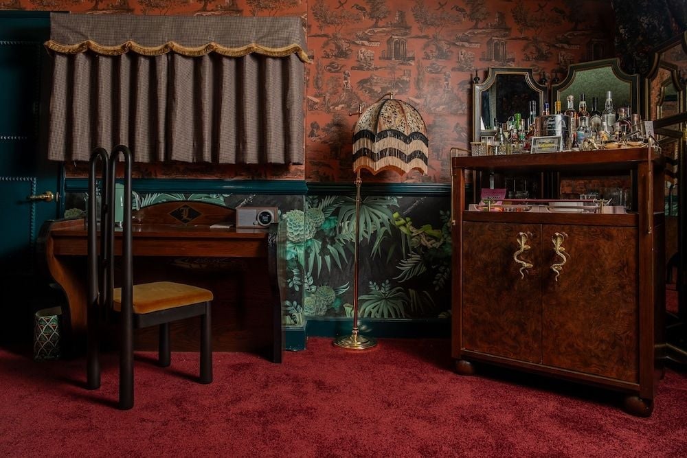 Plush burgundy carpeting and tropical wallpaper create an intimate speakeasy atmosphere, where vintage brass details and rich wood furnishings invite lingering conversations over craft cocktails.