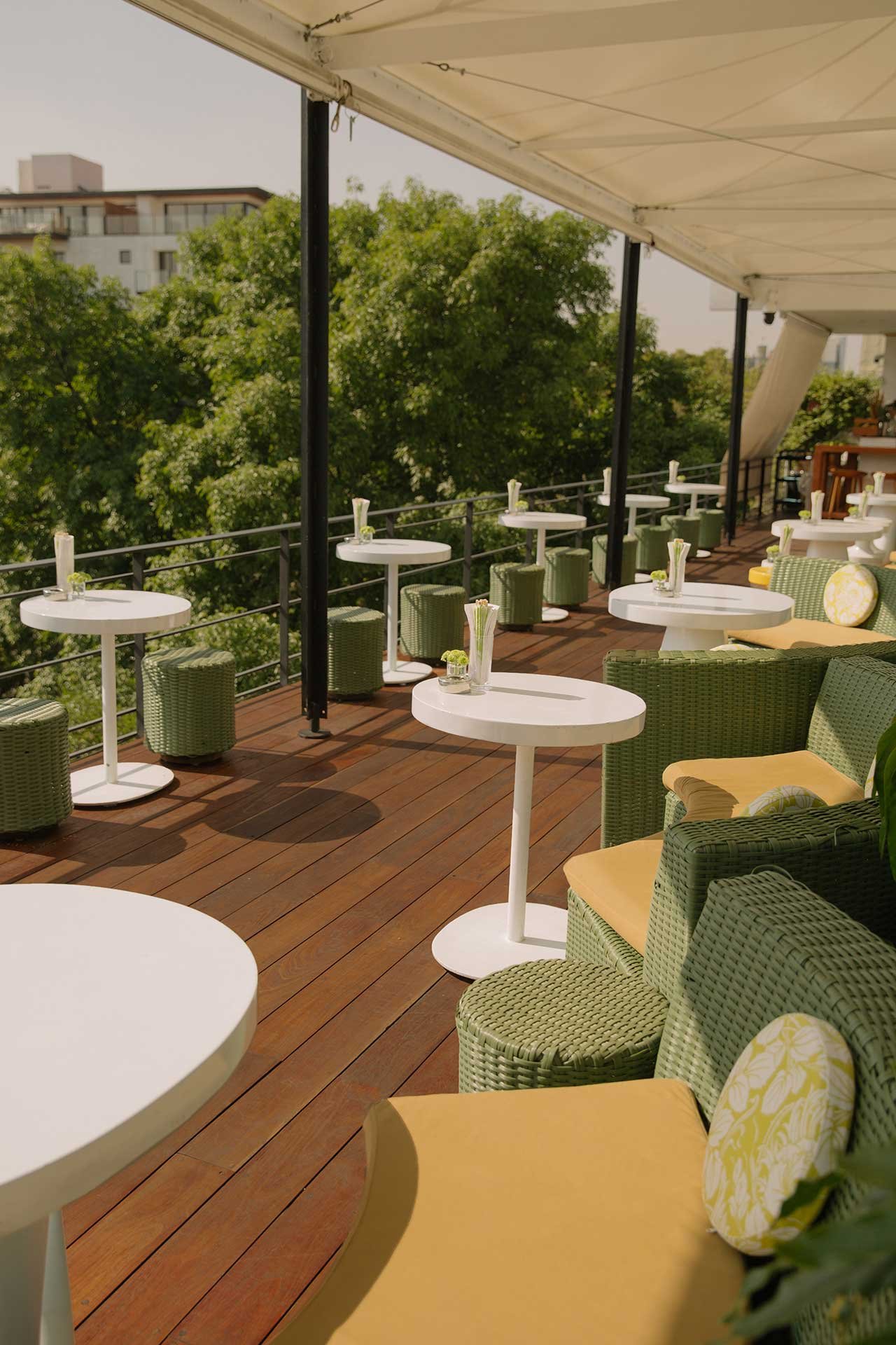 Green wicker chairs and crisp white cocktail tables create an intimate rooftop retreat, where canopied shade frames Mexico City's leafy skyline through the afternoon haze.