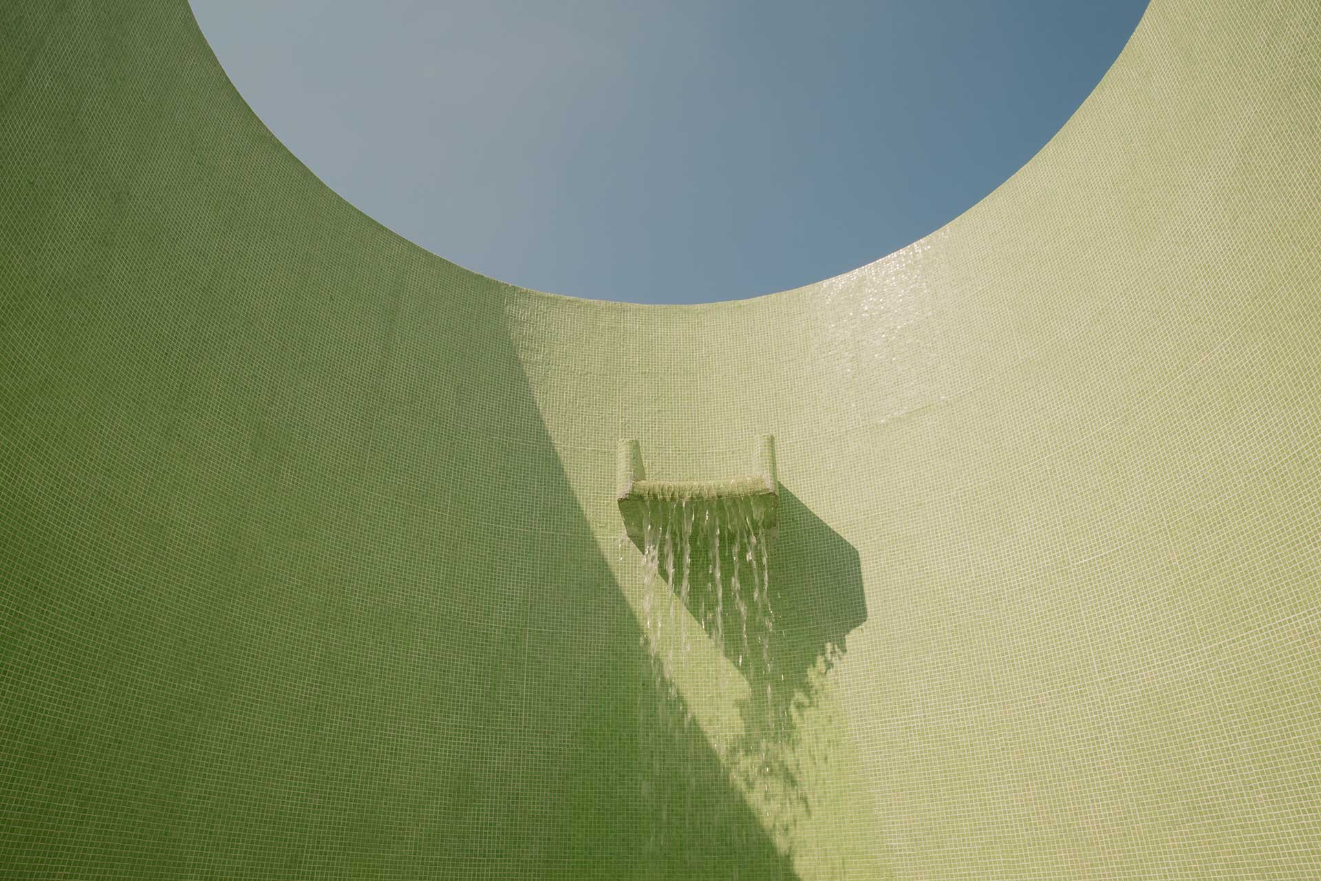 Water cascades from a sculptural spout into the rooftop pool, creating gentle ripples across the surface while geometric shadows play on the surrounding deck.