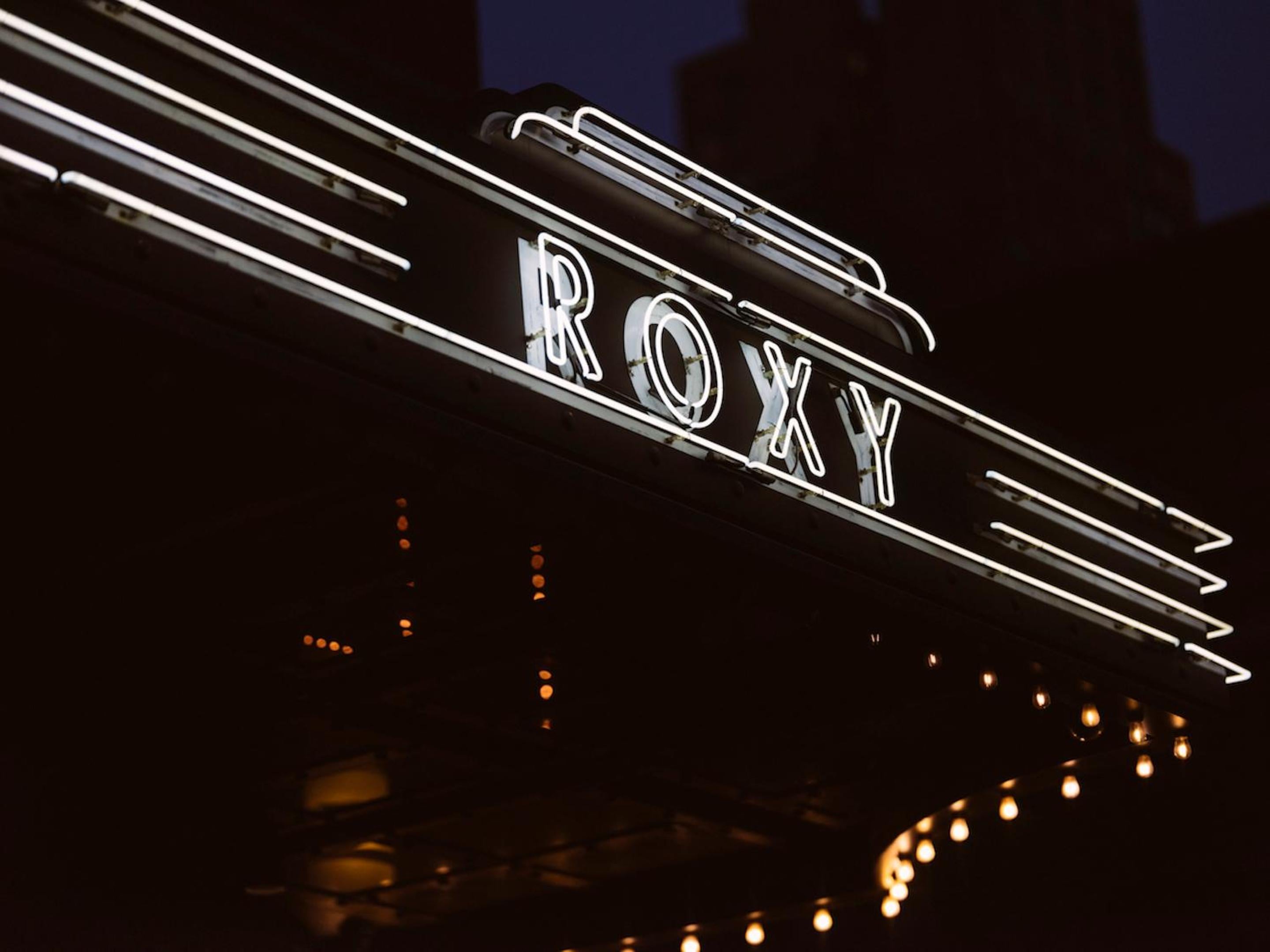 Art Deco-inspired neon lettering illuminates the Roxy's theatrical facade, where white light tubes create dramatic shadows against the evening sky in classic New York style.