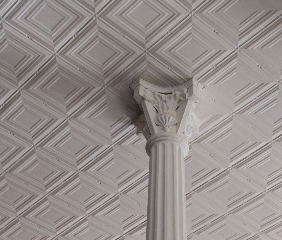Ornate tin ceiling tiles frame a classical column's Corinthian capital, where heritage architecture meets contemporary design in this thoughtfully restored heritage space.