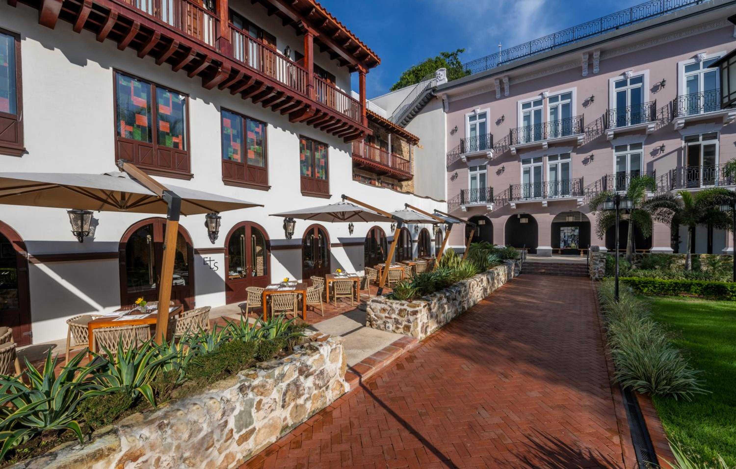 Sunlight filters through umbrellas onto the brick courtyard where colonial arches frame intimate dining terraces, while colorful window details peek from weathered balconies above.