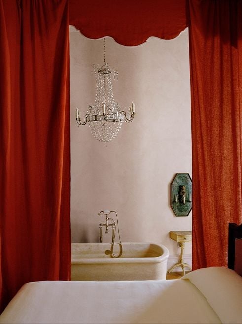 Crimson silk curtains frame an elegant soaking tub beneath a crystal chandelier, where vintage brass fixtures and classical details create an unexpectedly theatrical bathing sanctuary.