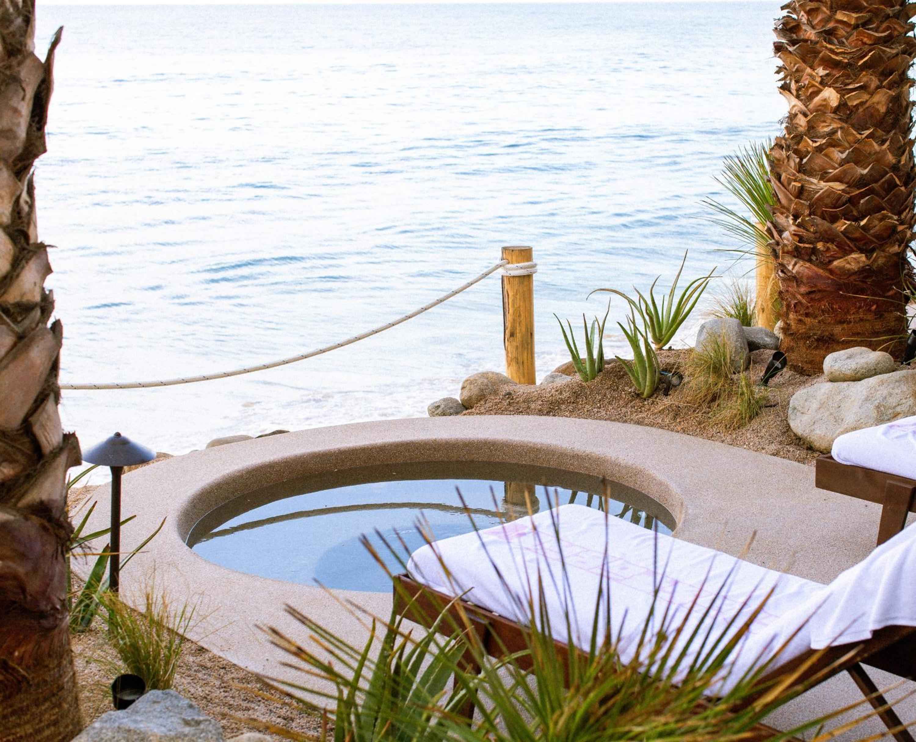 Sand-colored stone curves around an intimate plunge pool where desert meets sea, palm fronds catching golden light as waves whisper beyond the rope barrier.