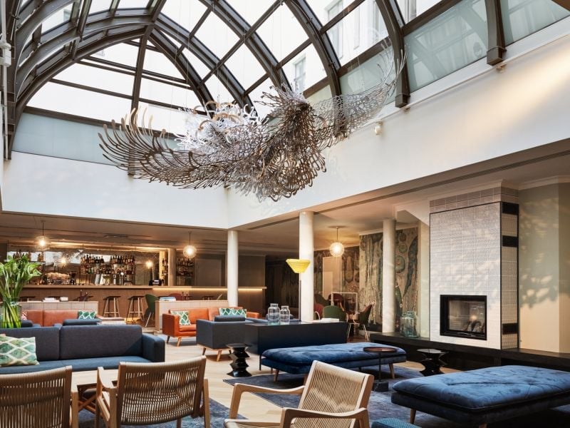 Soaring glass ceiling frames an intricate wire sculpture above plush navy seating, where contemporary design meets artistic sophistication in this stunning atrium lounge.