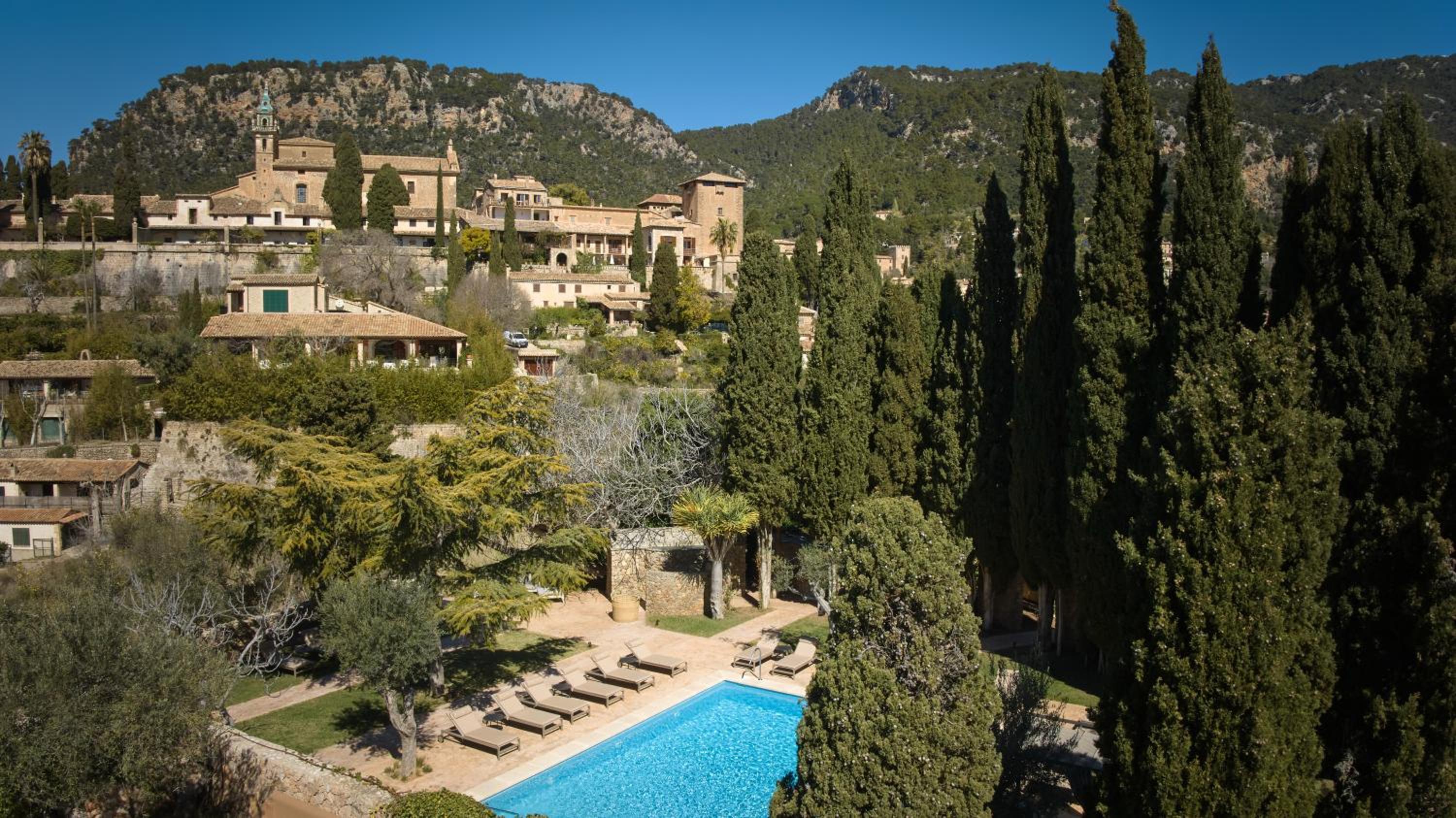 Stone terraces cascade down the hillside where cypress trees frame a pristine pool, while the ancient Mallorcan village of Valldemossa rises majestically beyond.