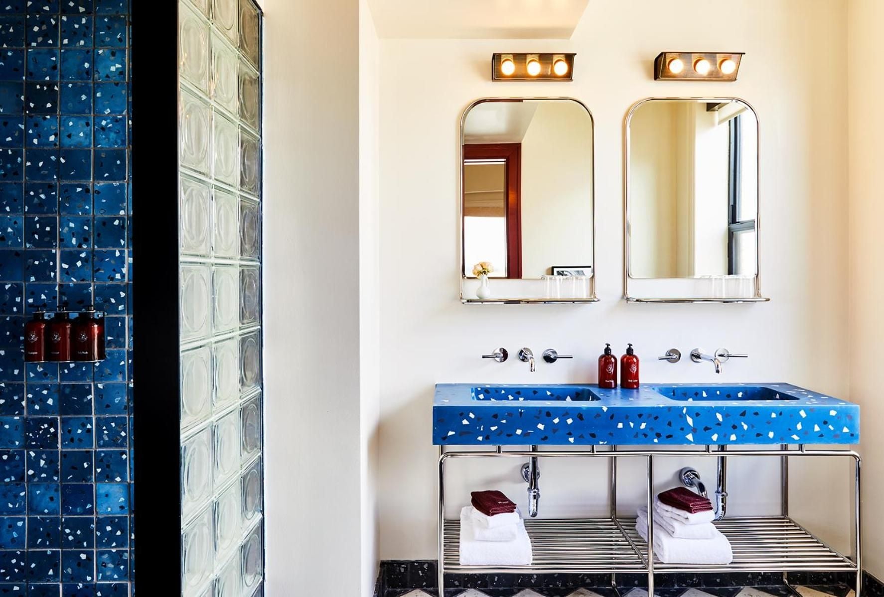 Vibrant blue terrazzo vanities with white speckled patterns create a striking focal point, while chrome fixtures and rounded mirrors add mid-century sophistication to this dual-sink sanctuary.