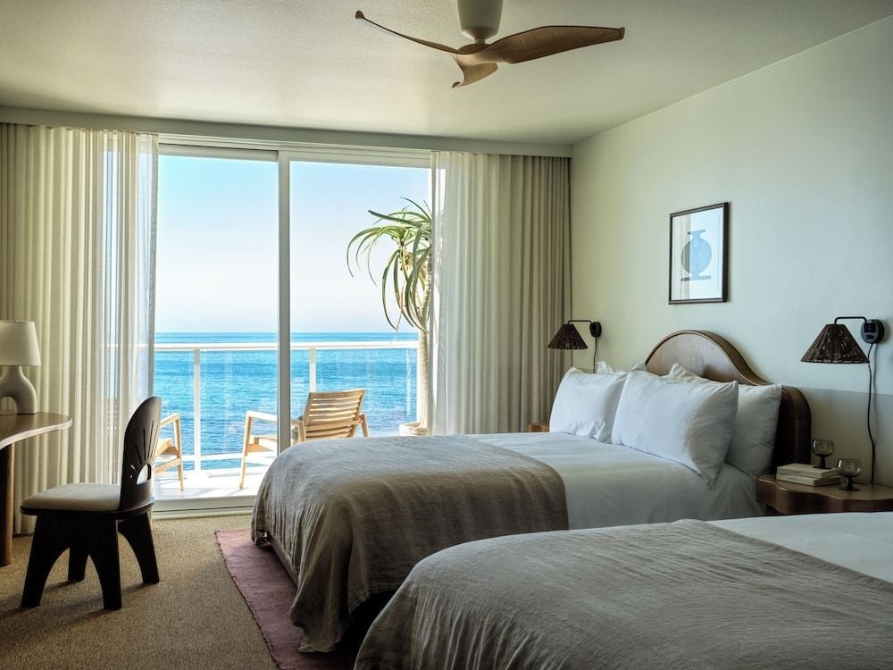 Twin beds frame floor-to-ceiling windows where ocean blues meet sky, while warm wood tones and flowing curtains create an airy coastal sanctuary above the waves.