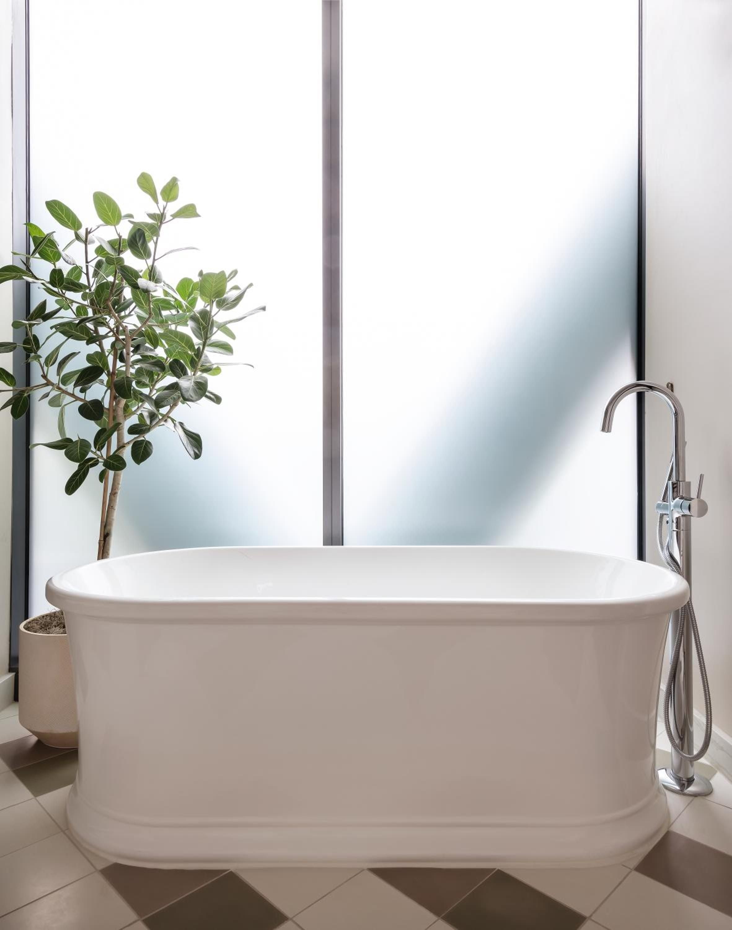 Sunlight streams through floor-to-ceiling windows, illuminating a sculptural soaking tub where modern minimalism meets spa-like serenity, complete with leafy greenery for natural warmth.