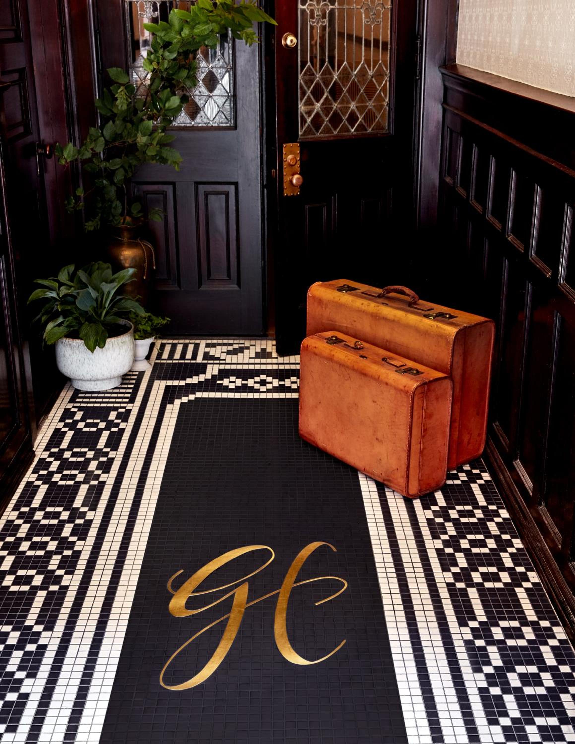 Vintage leather suitcases rest on intricate mosaic tiles in Guild House's sophisticated entryway, where custom monogramming and rich mahogany paneling evoke timeless elegance.