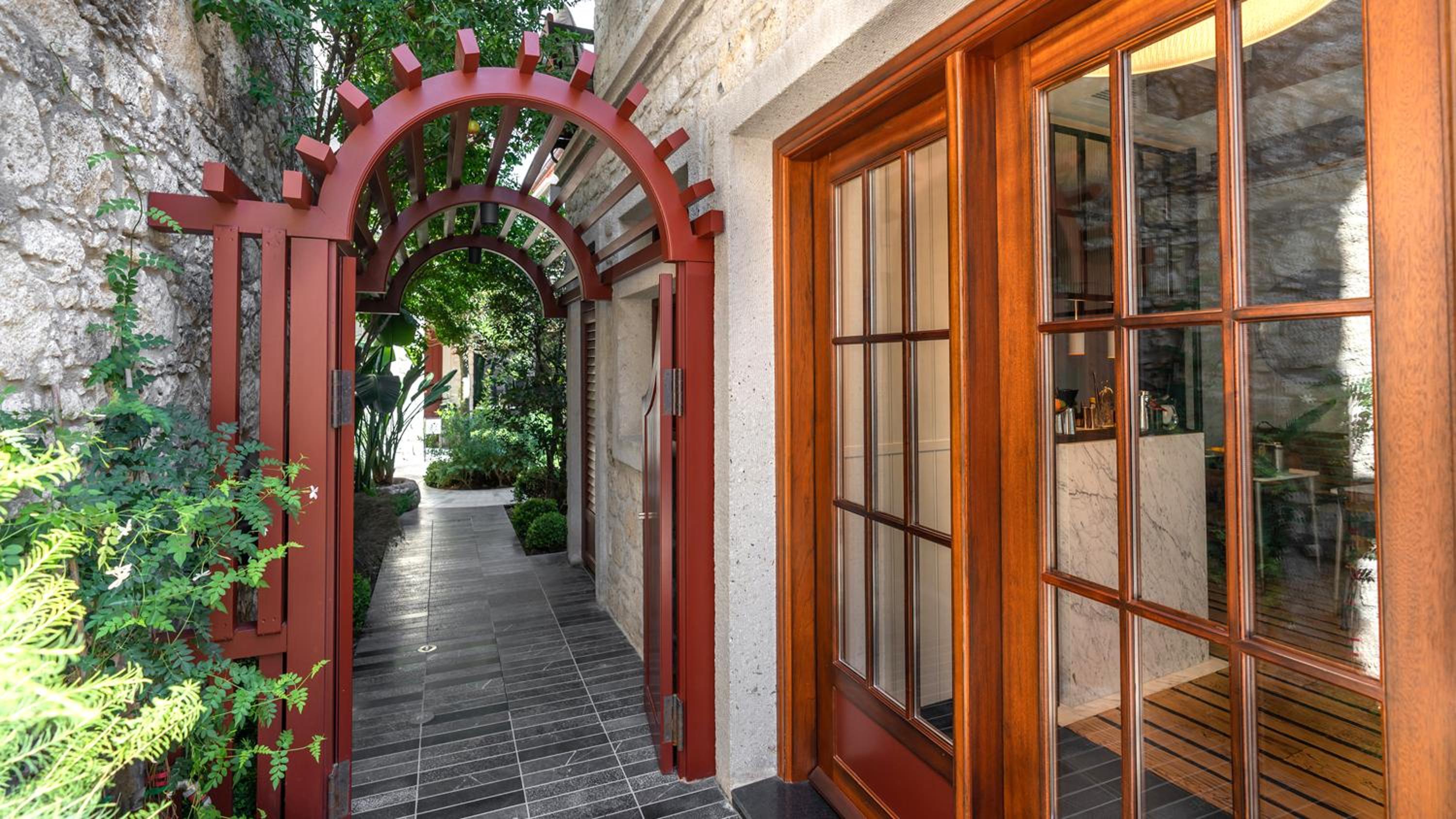 Weathered stone walls frame an inviting red pergola gateway, where dappled sunlight filters through lush greenery onto gleaming wood floors beyond.