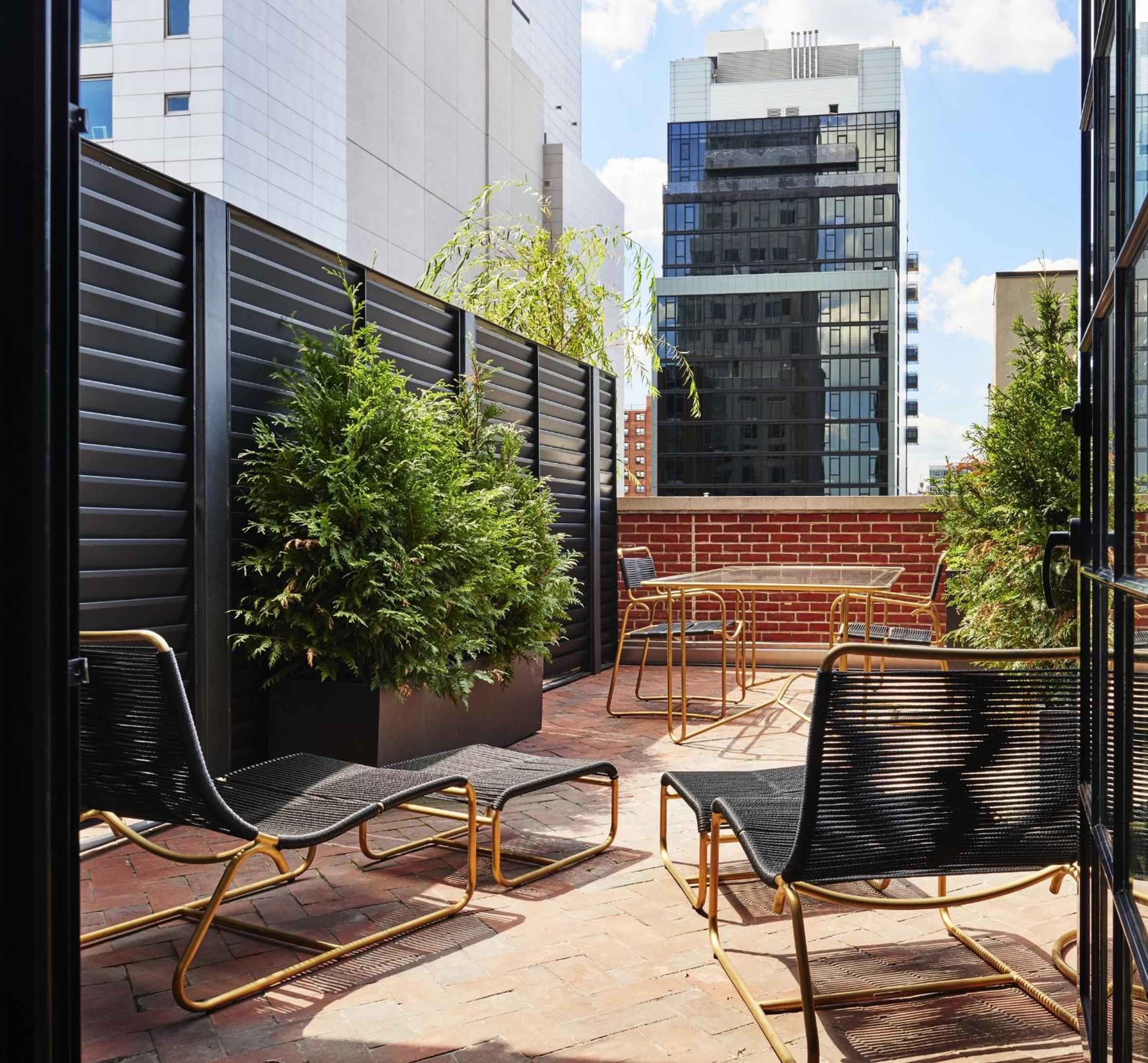 Golden-framed lounge chairs catch afternoon light on this brick-walled terrace, where privacy screens frame glimpses of Manhattan's towering glass and steel beyond.