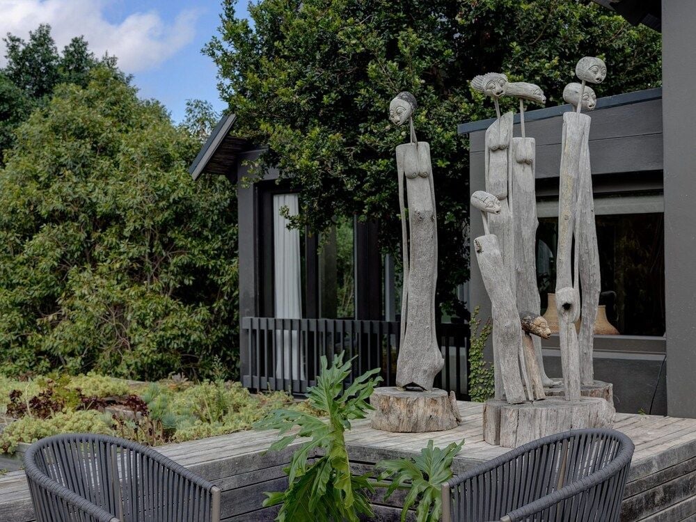 Weathered driftwood sculptures rise like silent sentinels on the wooden deck, their organic forms creating intimate conversation nooks amid lush plantings and modern architecture.