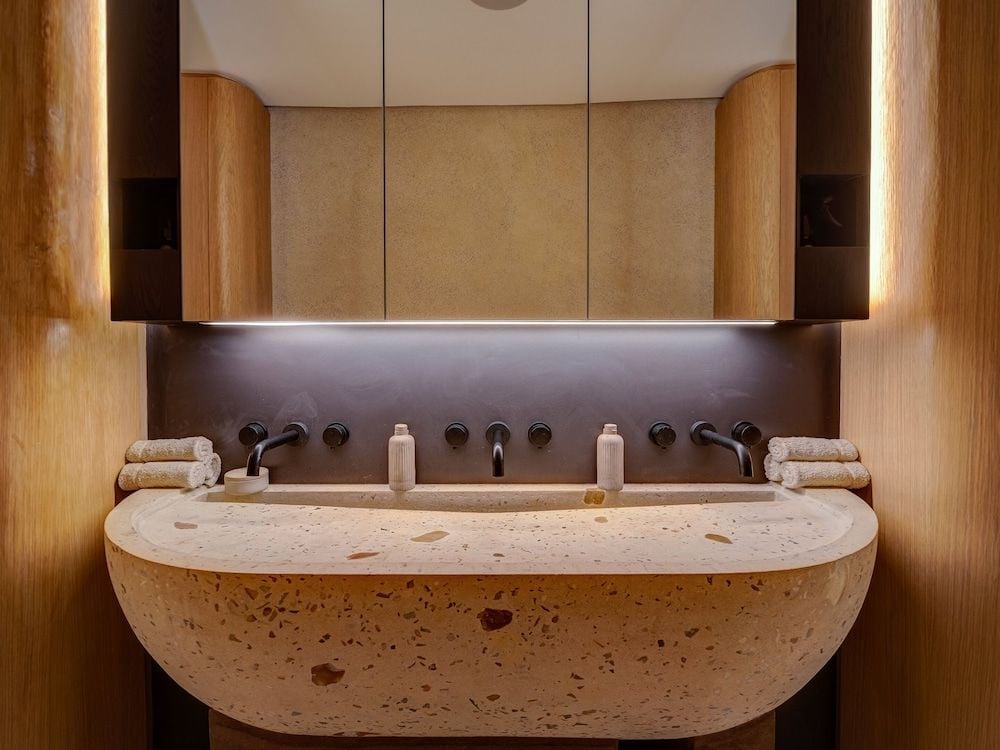 Curved terrazzo basins and warm wood columns create an intimate double vanity where under-cabinet lighting casts a gentle glow across speckled stone surfaces.