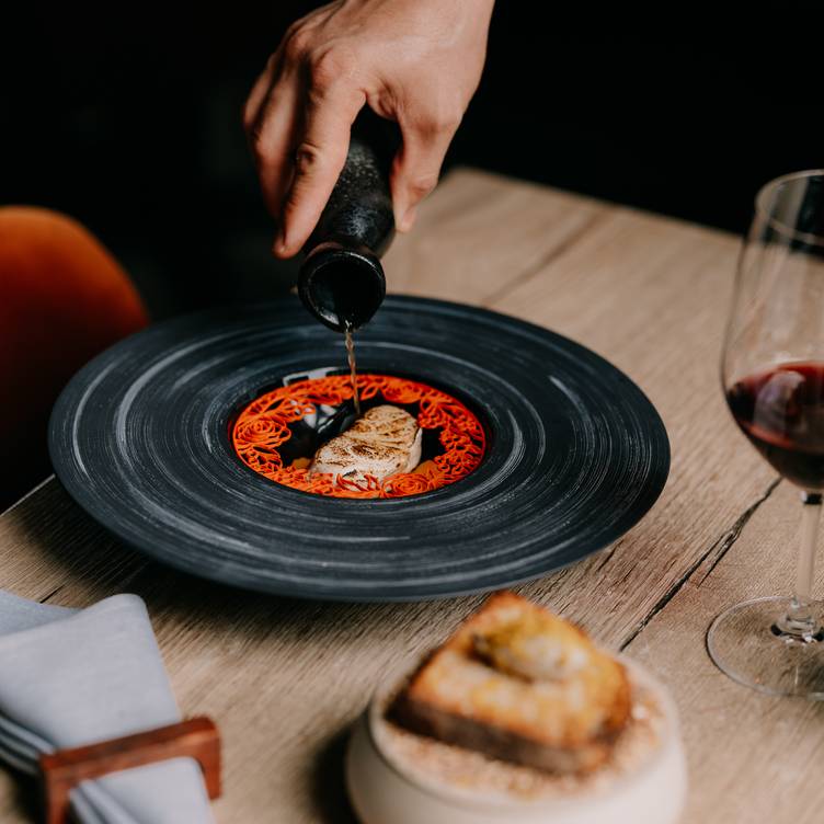 A hand drizzles dark sauce onto artfully plated foie gras, transforming the charcoal-rimmed dish into edible art alongside crusty bread and wine.