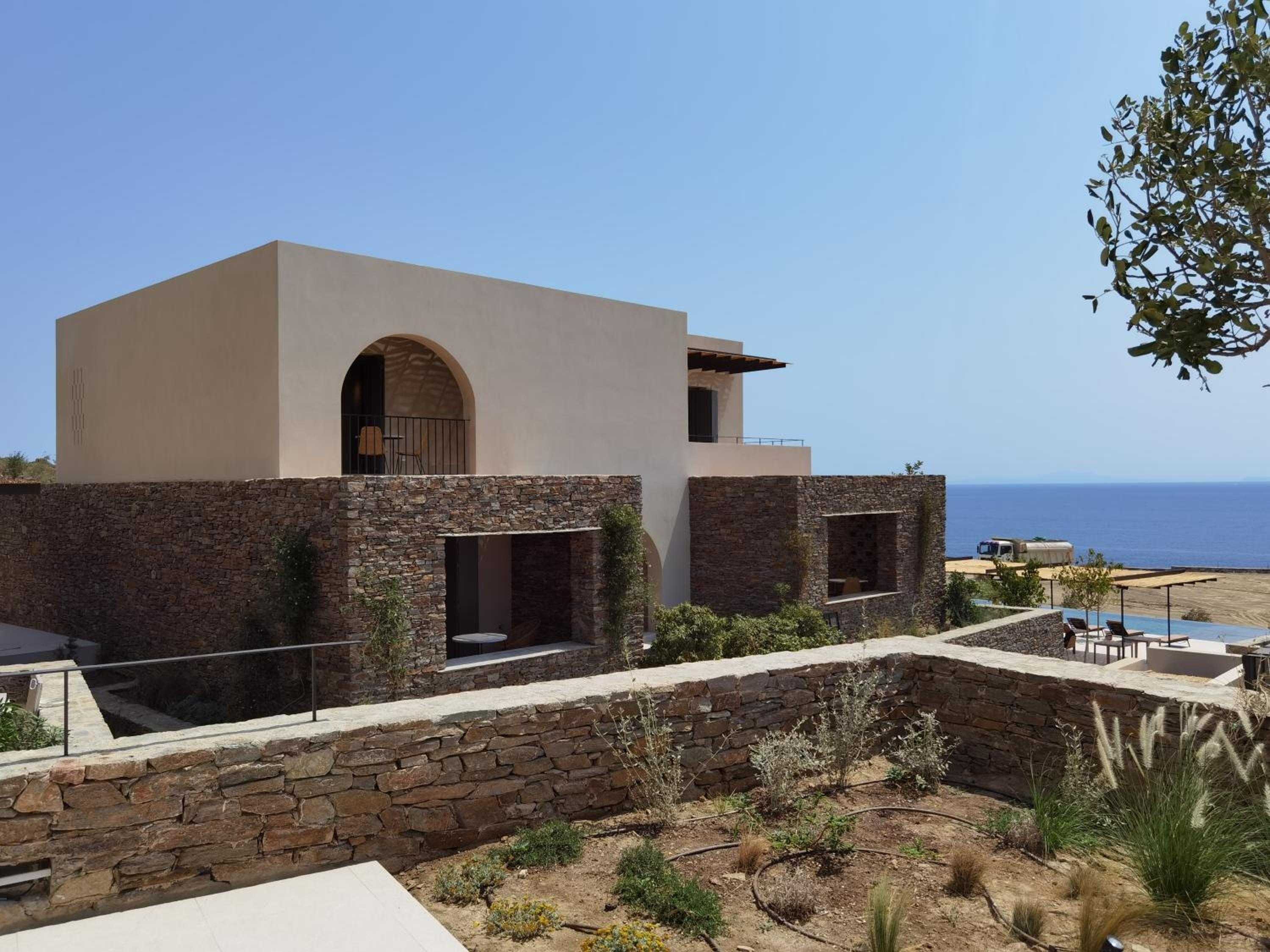 Stone-wrapped terraces cascade down the clifftop, where contemporary cube architecture meets ancient Cycladic masonry against the endless Aegean blue.