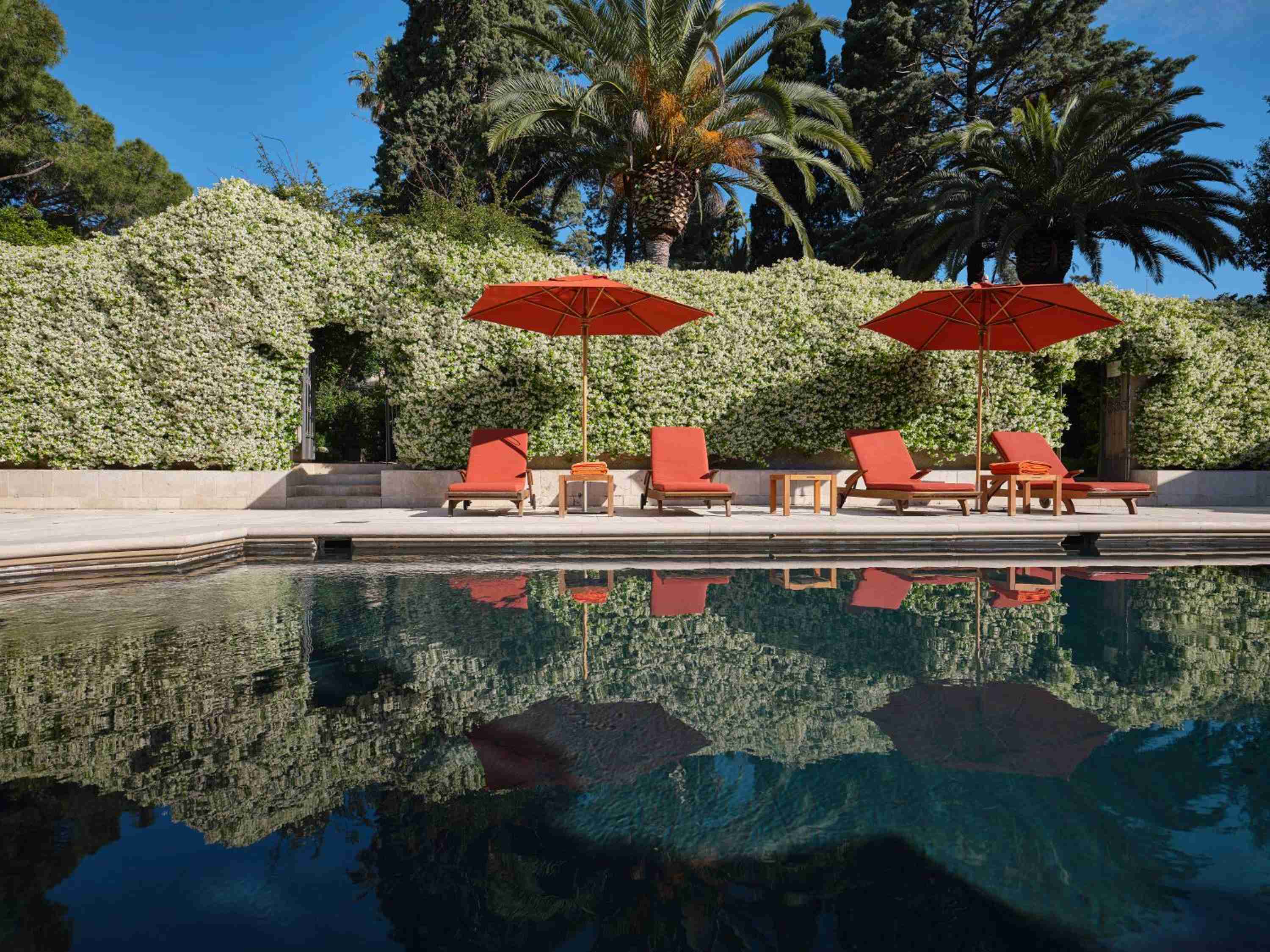 Orange loungers line the infinity pool beneath swaying palms and jasmine-covered walls, their vibrant reflections dancing on crystalline water under Basilicata's azure sky.