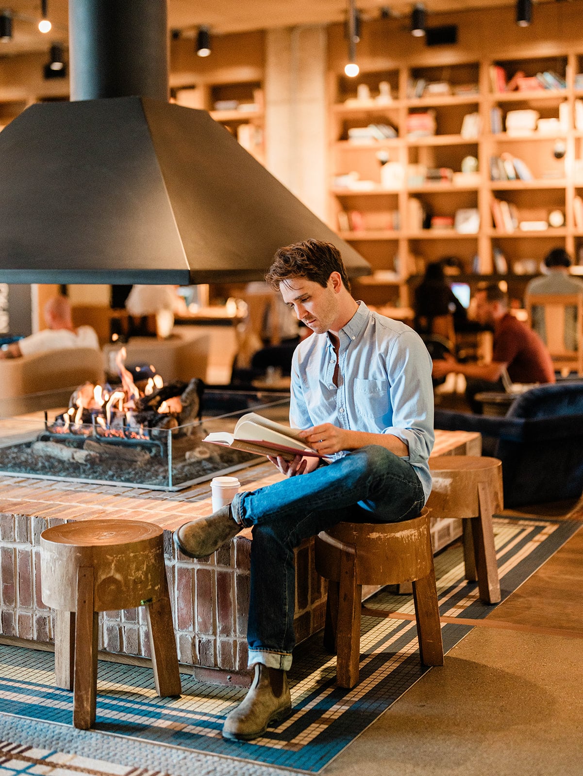 Crackling flames dance in the modern fireplace as guests settle into rustic wooden stools with books and coffee, surrounded by towering shelves of curated reads.