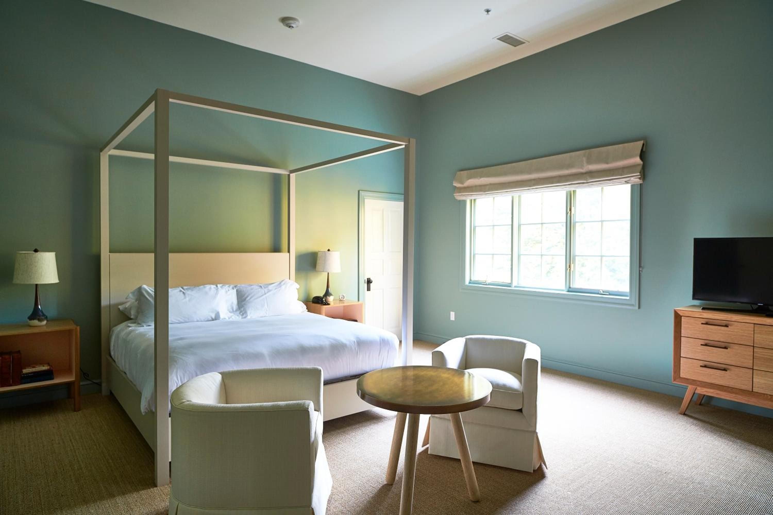 Sage-green walls and a sleek four-poster frame create serene sanctuary vibes, while mid-century furnishings and crisp white linens invite lingering mornings in the Hudson Valley.