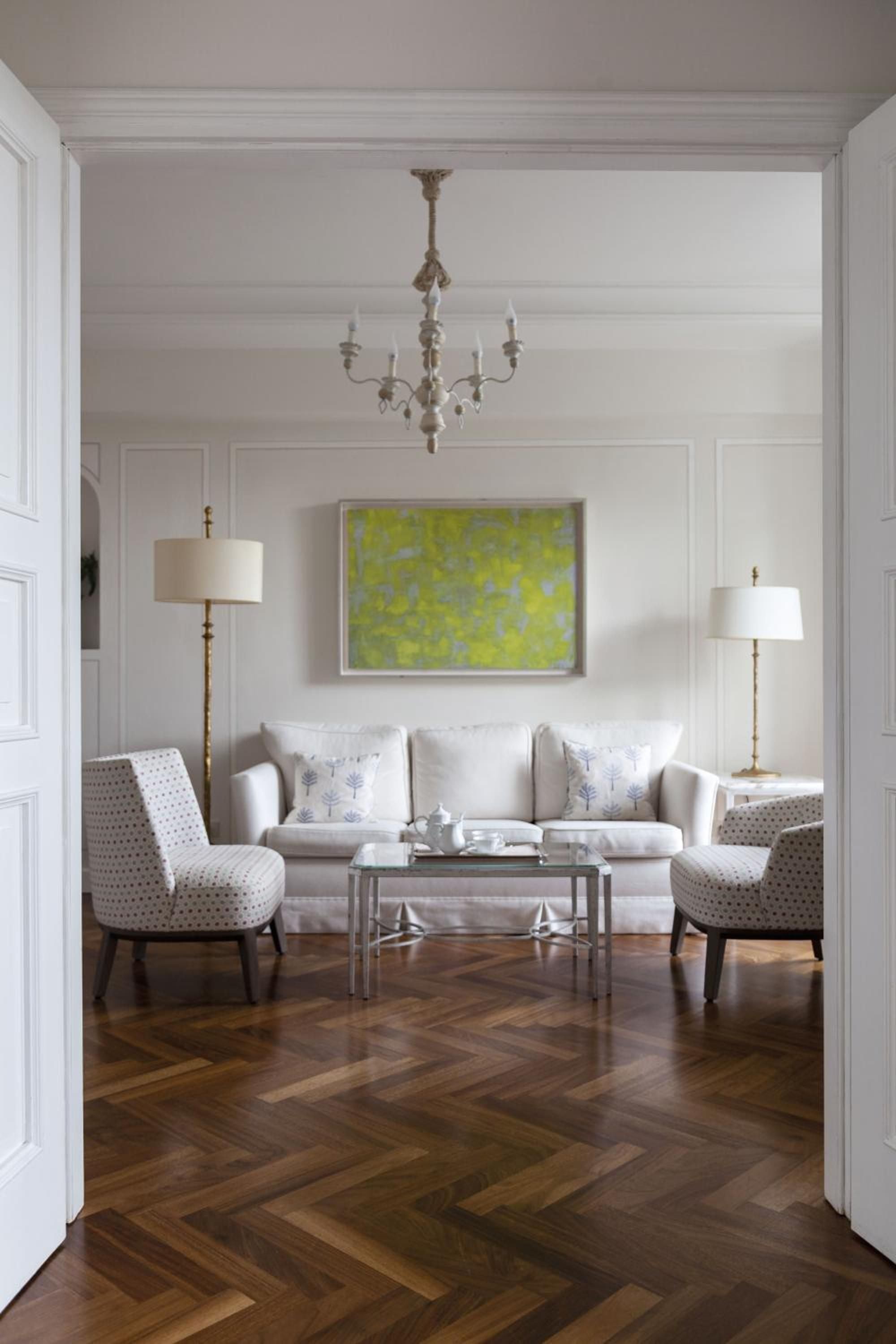 Soft morning light filters through this elegant sitting area, where polished herringbone floors meet crisp white furnishings and a vibrant chartreuse artwork adds unexpected Mediterranean flair.