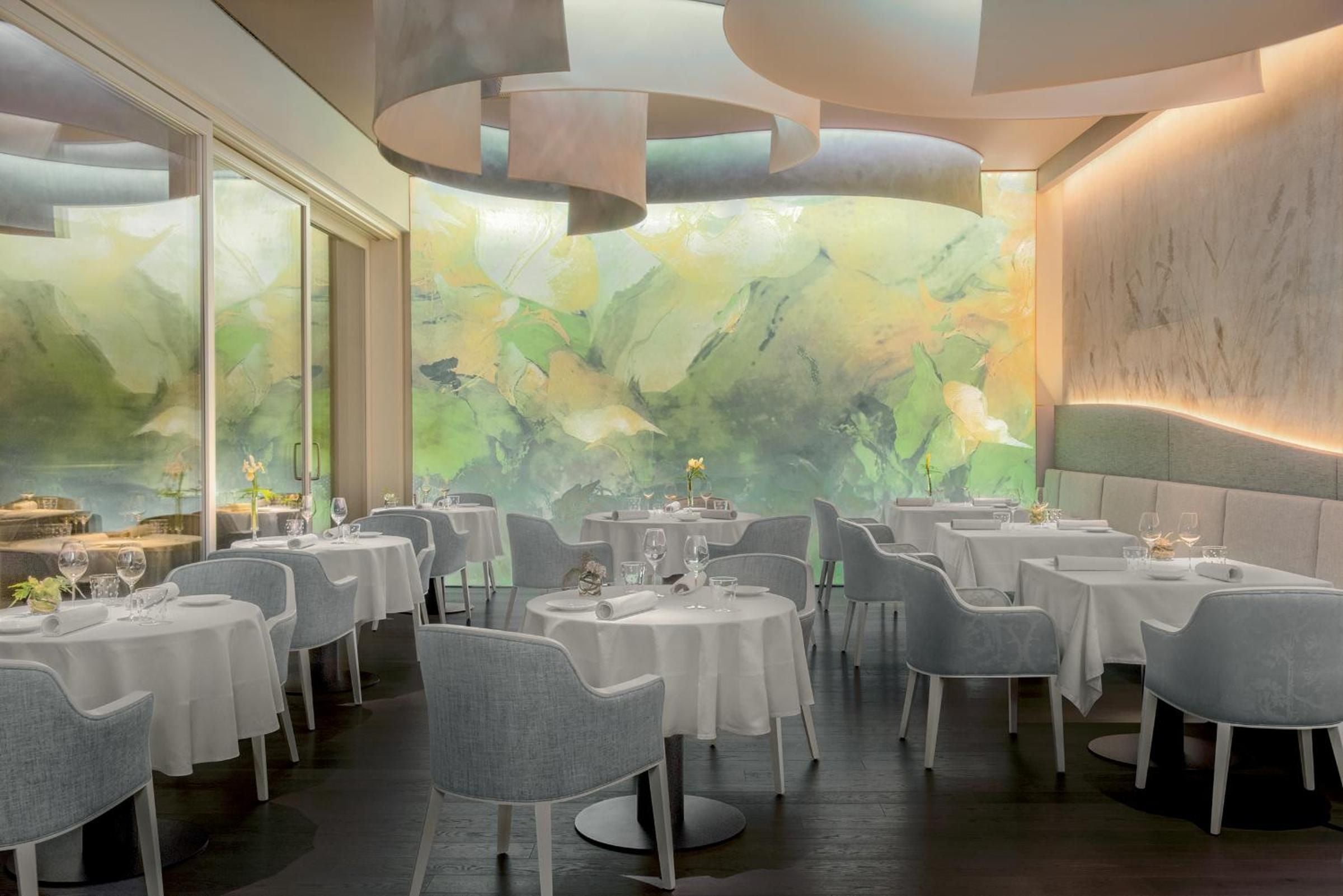 Sophisticated dining unfolds beneath sculptural ceiling details, where sage-toned chairs face floor-to-ceiling windows revealing impressionistic garden murals that blur indoor-outdoor boundaries.