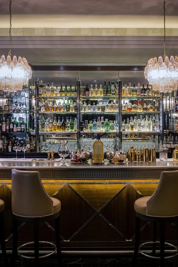 Crystal chandeliers cast warm light over the sleek marble bar, where hundreds of premium spirits gleam on illuminated shelves behind plush leather stools.