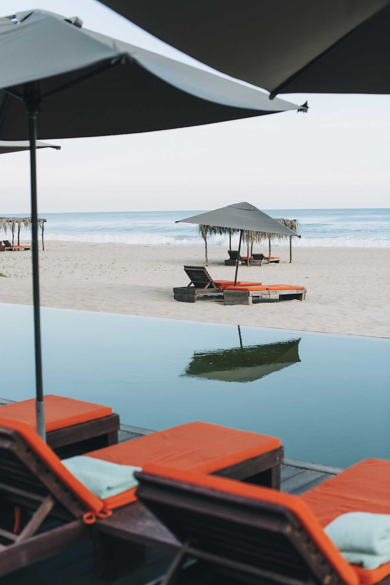 Orange-cushioned loungers frame the infinity pool where calm waters mirror passing boats, while thatched umbrellas dot the pristine shoreline beyond.