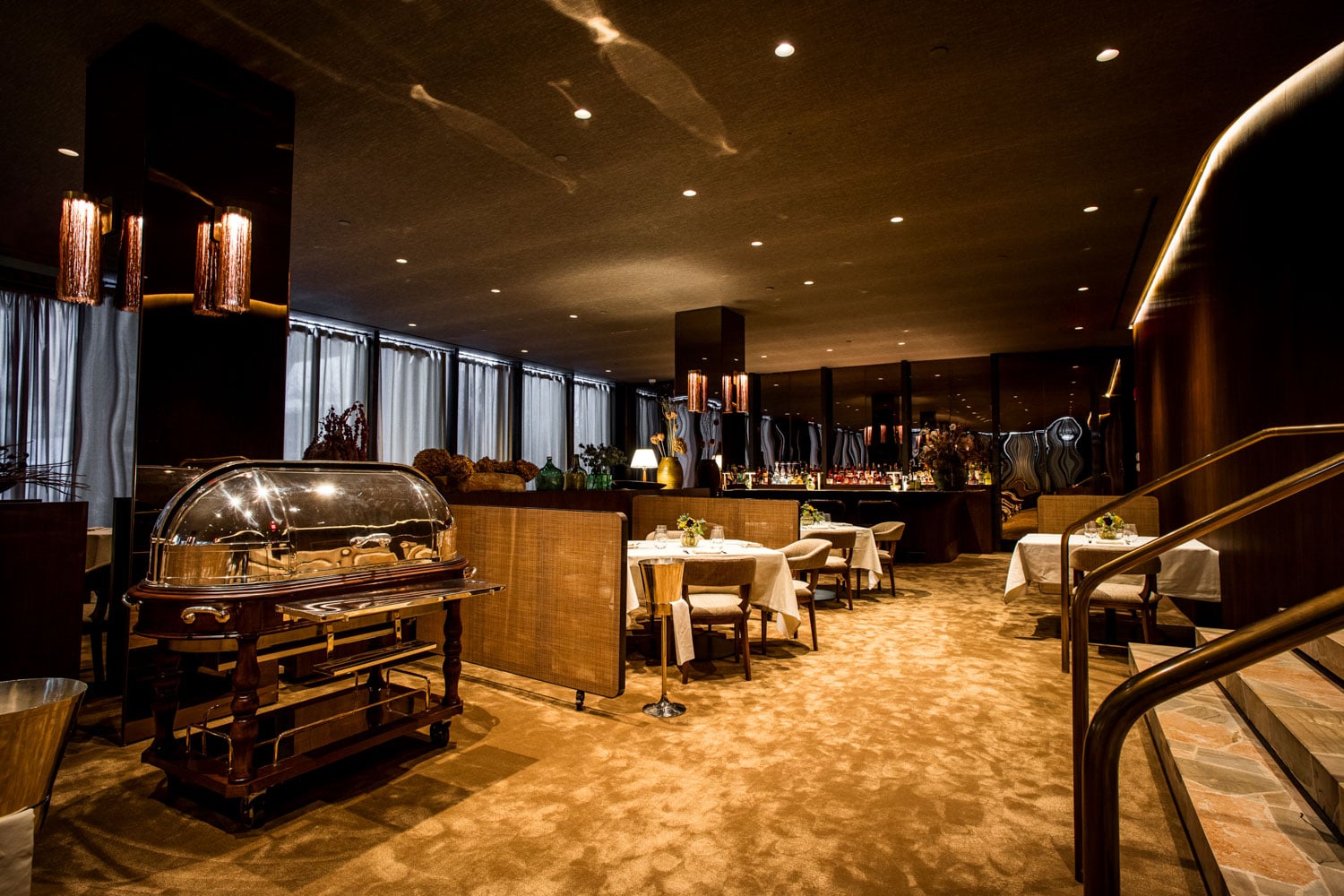 Copper pendant lights cast a warm glow over polished serving carts and intimate tables, where rich textures and curved railings create an unmistakably sophisticated dining atmosphere.