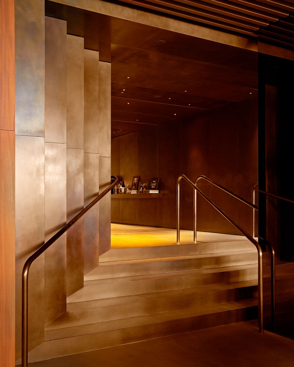 Warm wood and bronze handrails guide guests through the hotel's sculptural staircase, where amber lighting creates an intimate counterpoint to soaring limestone columns.