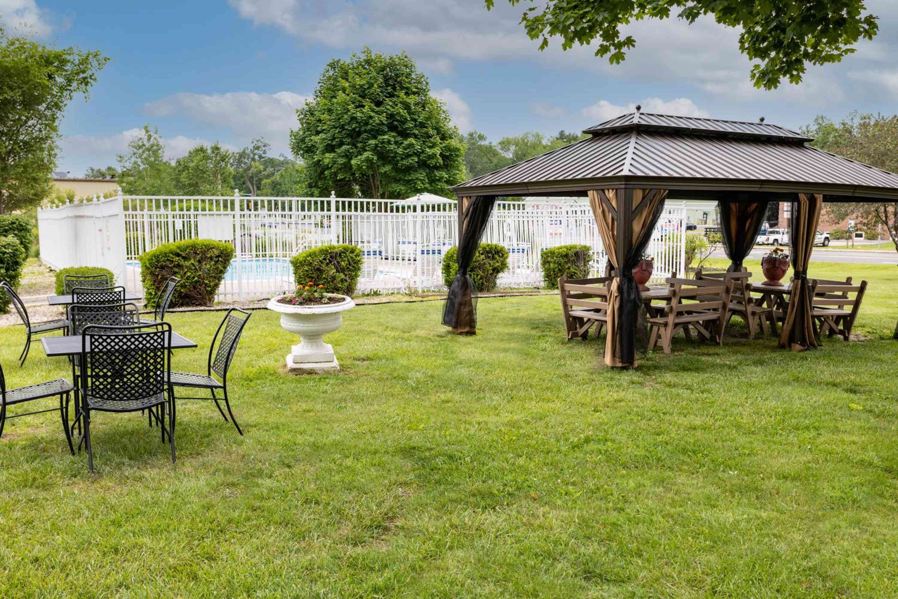 Manicured lawns stretch between wrought-iron seating and a rustic gazebo, where hanging planters and privacy curtains create intimate outdoor dining beneath towering shade trees.