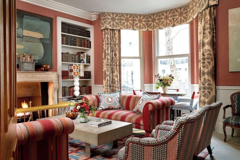 Rich coral walls and flame-patterned curtains create an intimate drawing room where leather-bound books and crackling fireplace invite lingering over afternoon tea.