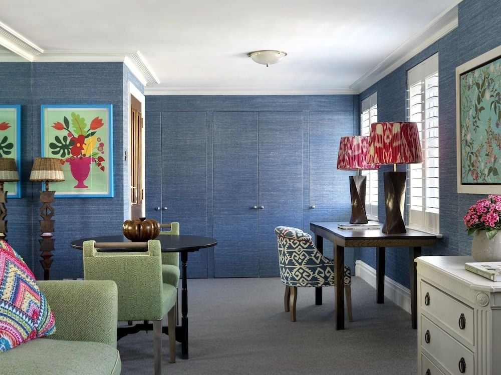 Sophisticated blue grasscloth wallpaper creates an intimate cocoon in this thoughtfully appointed guest room, where vibrant florals and geometric patterns add personality to classic furnishings.