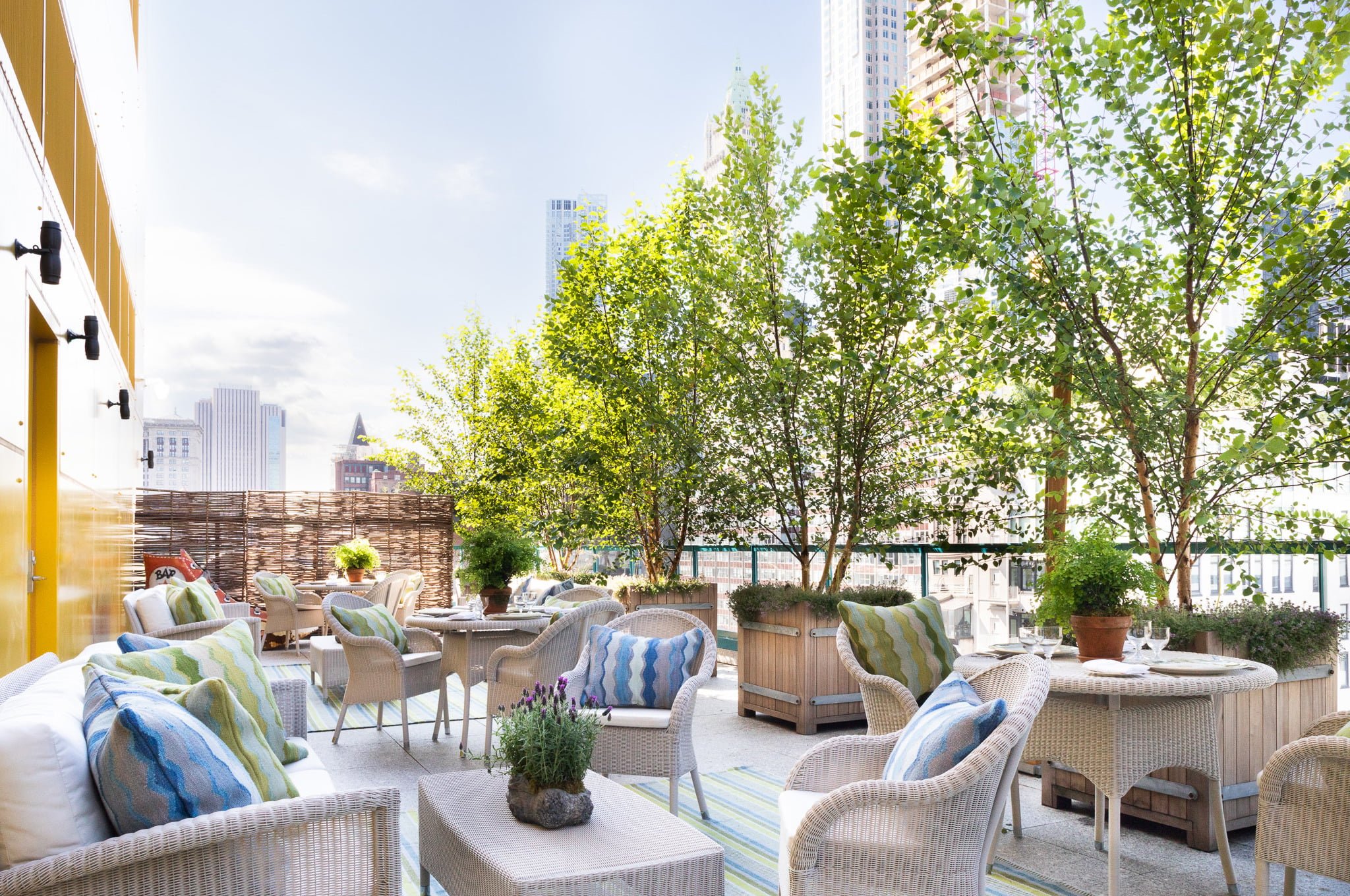 Wicker furnishings with cheerful blue-green cushions create intimate conversation nooks on this tree-lined rooftop terrace, where Manhattan's skyline provides a stunning urban backdrop.