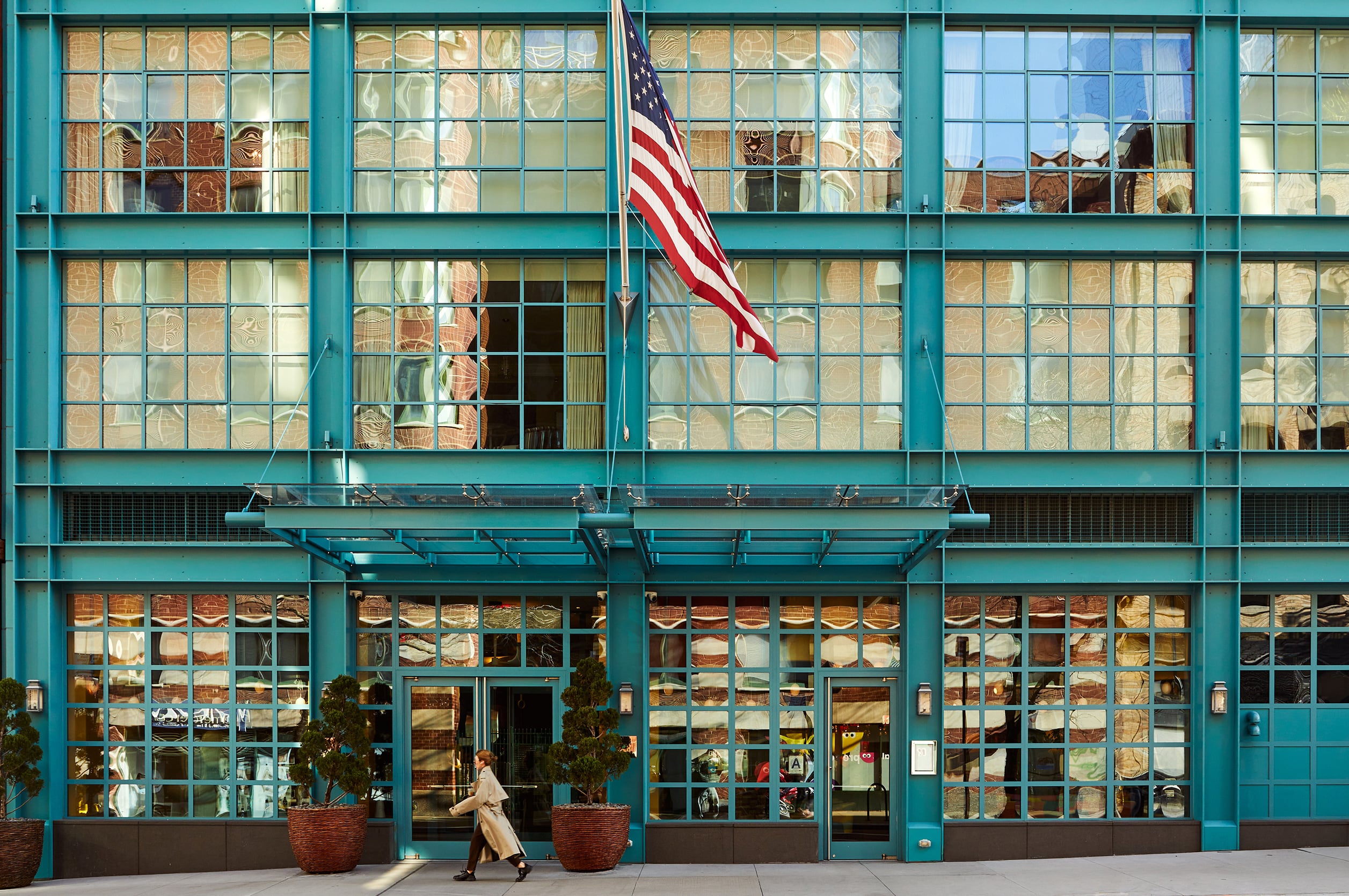 Turquoise steel frames create a striking grid pattern across the hotel's facade, where an American flag waves above the bustling street-level entrance filled with warm golden light.